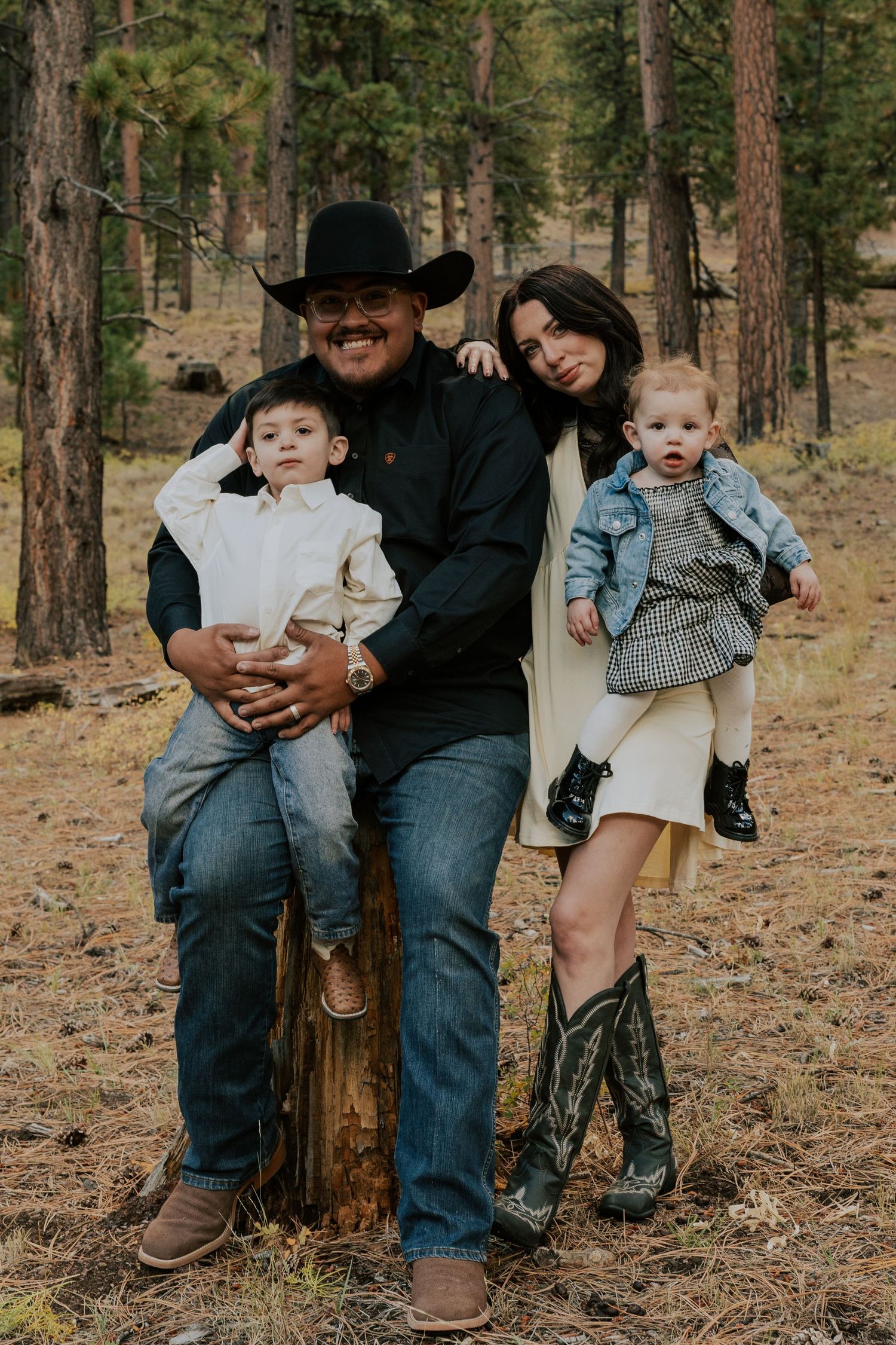 Family of four posing together by a tree stump in a pine forest, dad wearing a black cowboy hat.