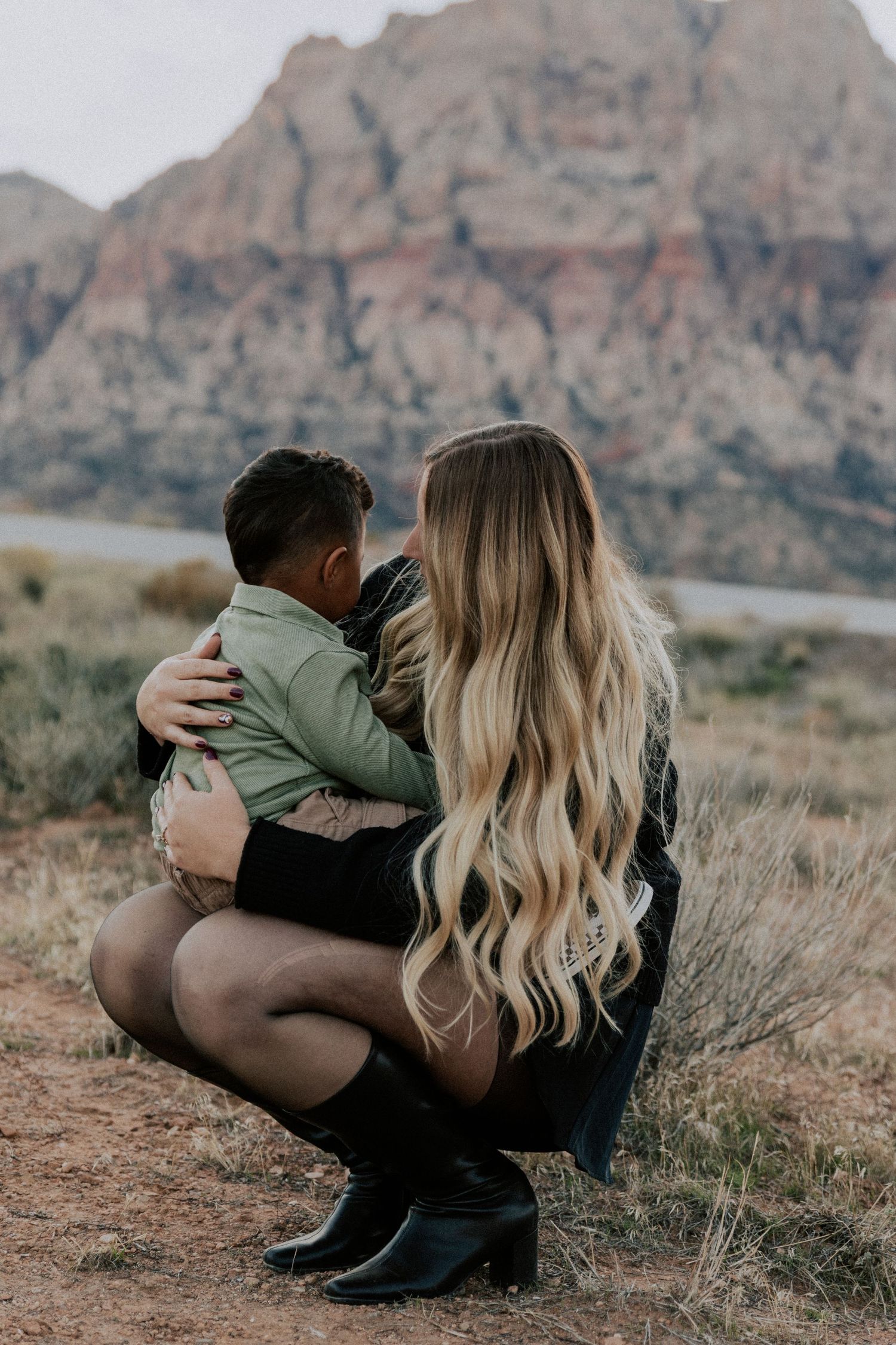 Woman with long blonde hair crouching to embrace a young child in a desert landscape with red rock mountains.