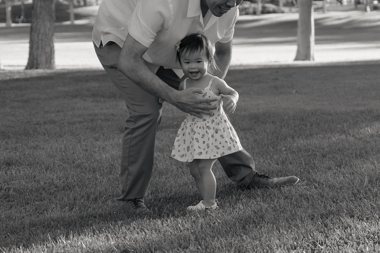 Black and white photo of grandfather helping smiling toddler girl walk on green grass in park.