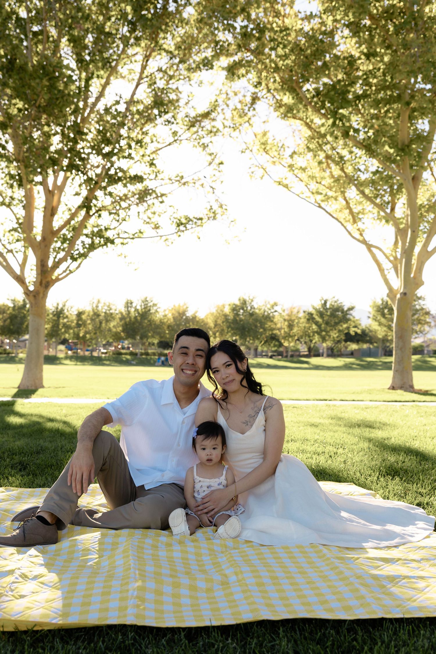 Young family with baby sitting on yellow checkered blanket in sunny park for engagement photos.
