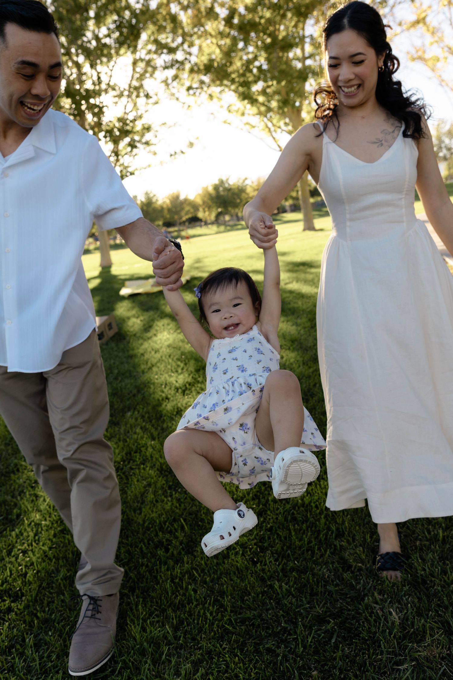 Joyful parents swinging laughing baby girl between them on lush green grass in sunny park.