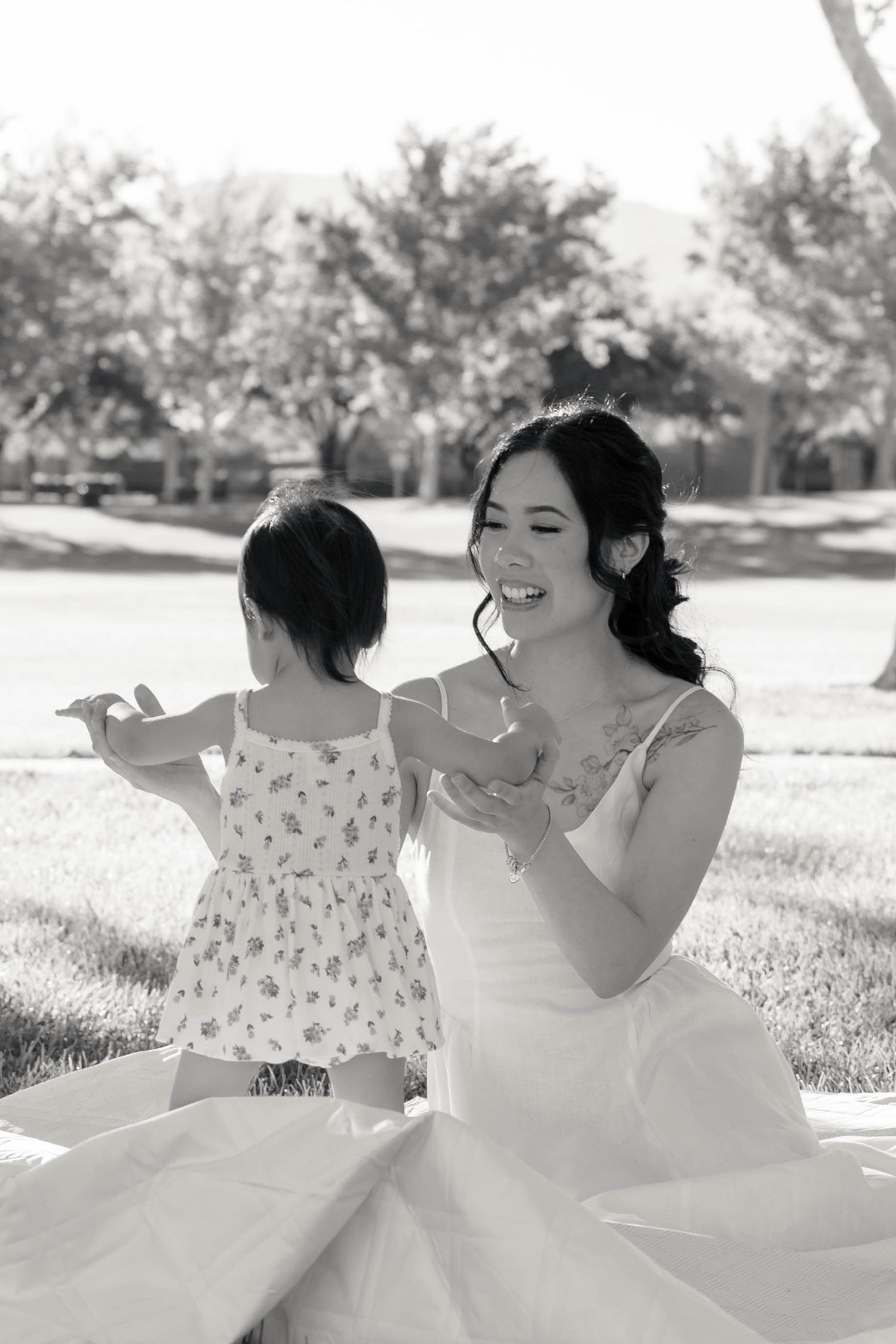 Black and white photo of smiling bride in white gown sitting on grass with young toddler outdoors.
