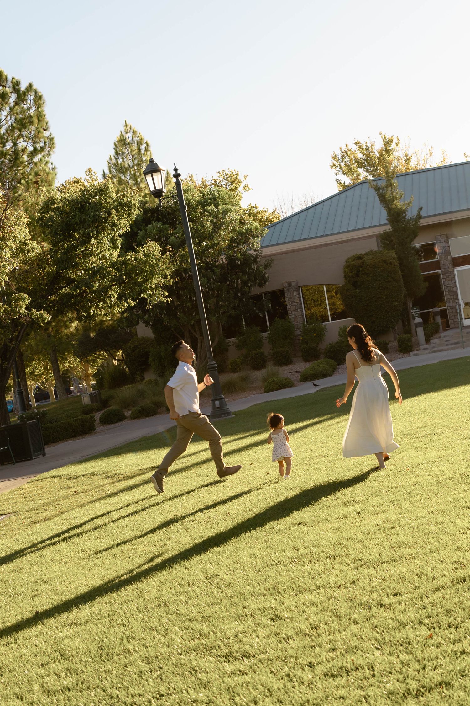 Young family running and playing together on green lawn in sunny park during golden hour session.