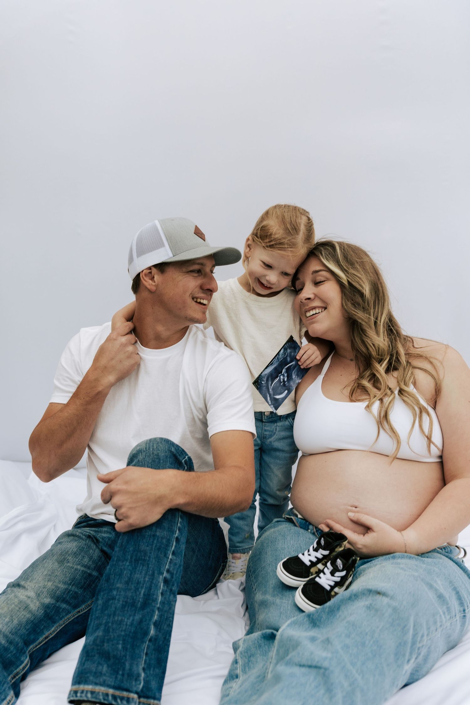 Smiling pregnant woman with husband and toddler son holding ultrasound photo on white background.