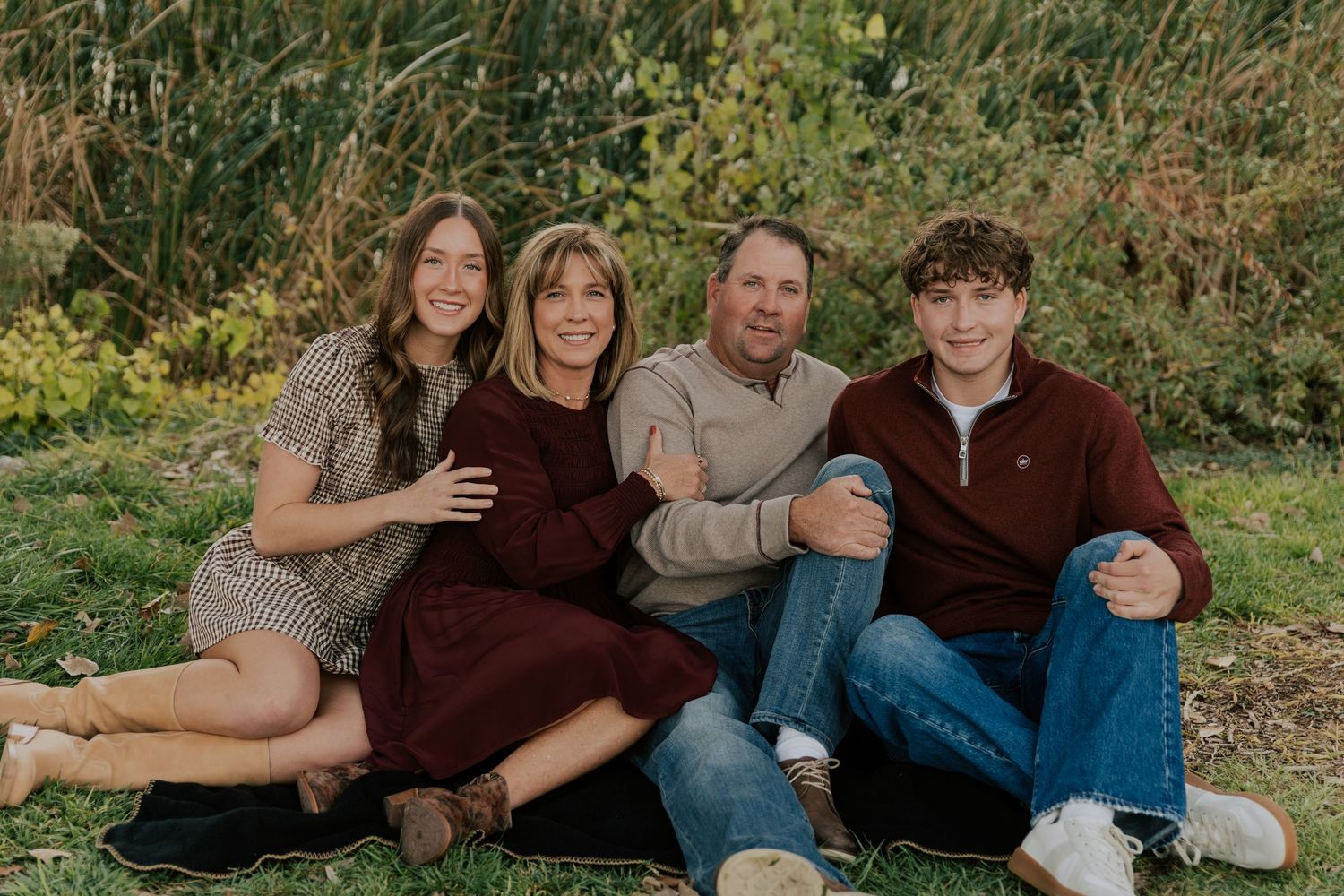 Family of four sitting together on green grass, smiling warmly in a cozy outdoor portrait session.