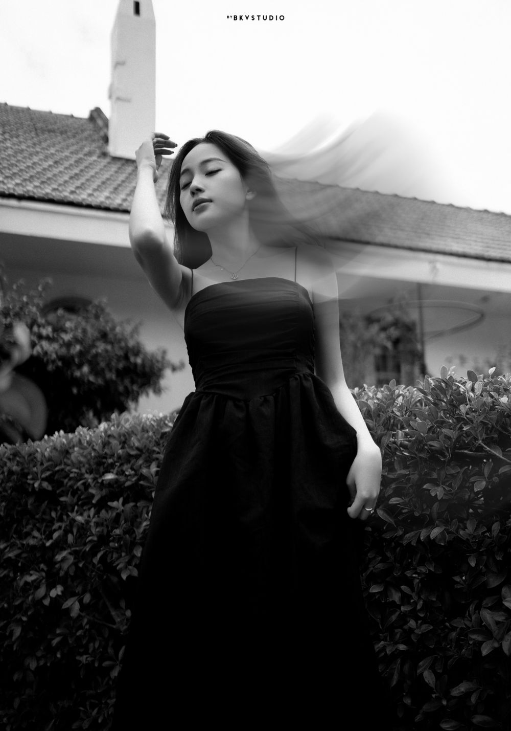 Artistic black and white portrait of a woman in a strapless black dress posing against lush garden foliage.