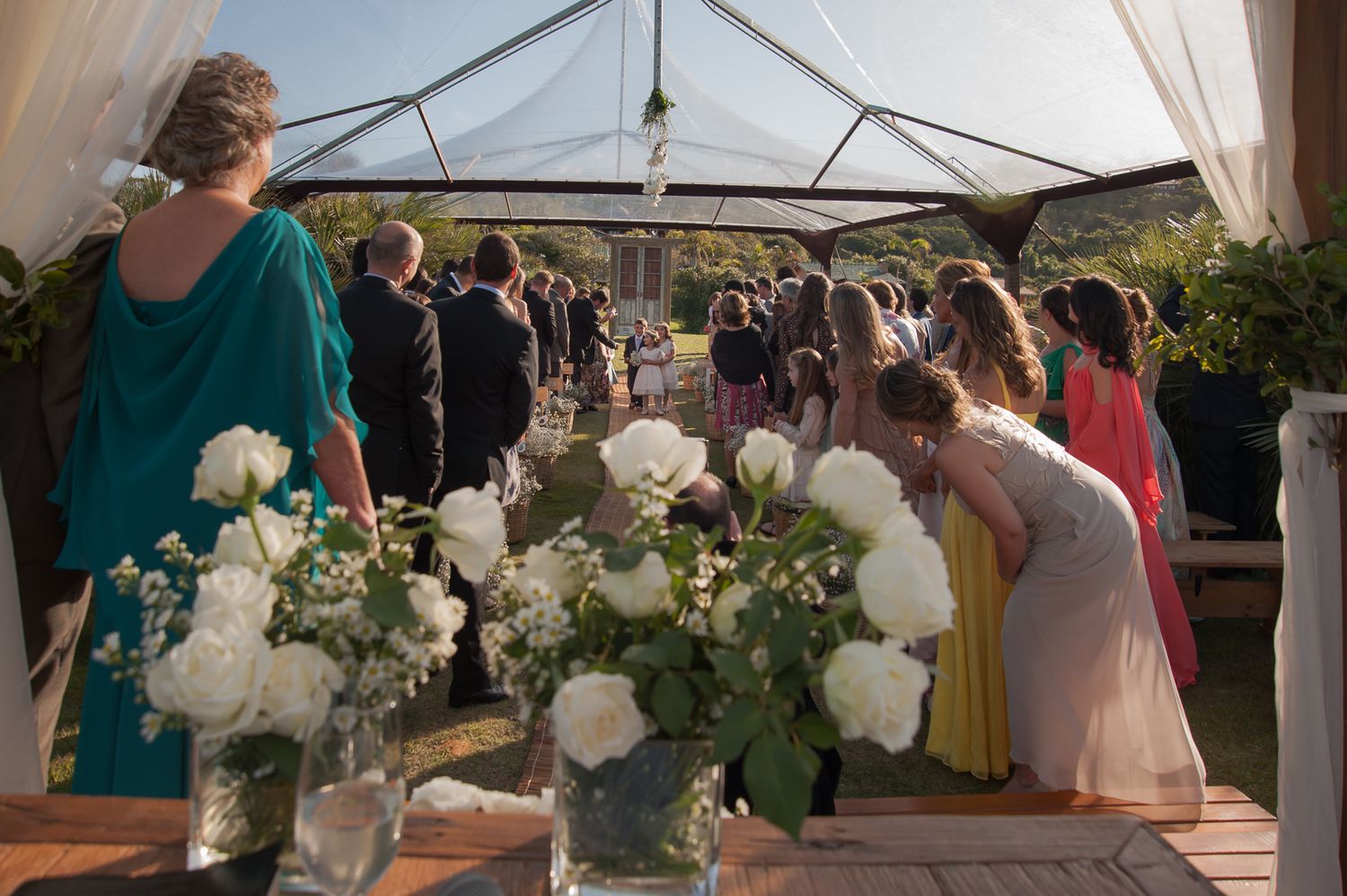 Wedding ceremony setup under white tent with floral arrangements and gathered guests.
