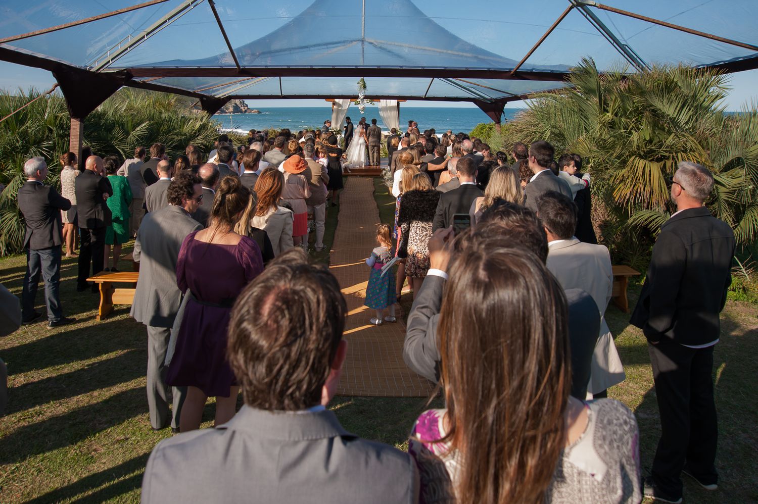 Wedding guests gather under a tent canopy for an outdoor ceremony.