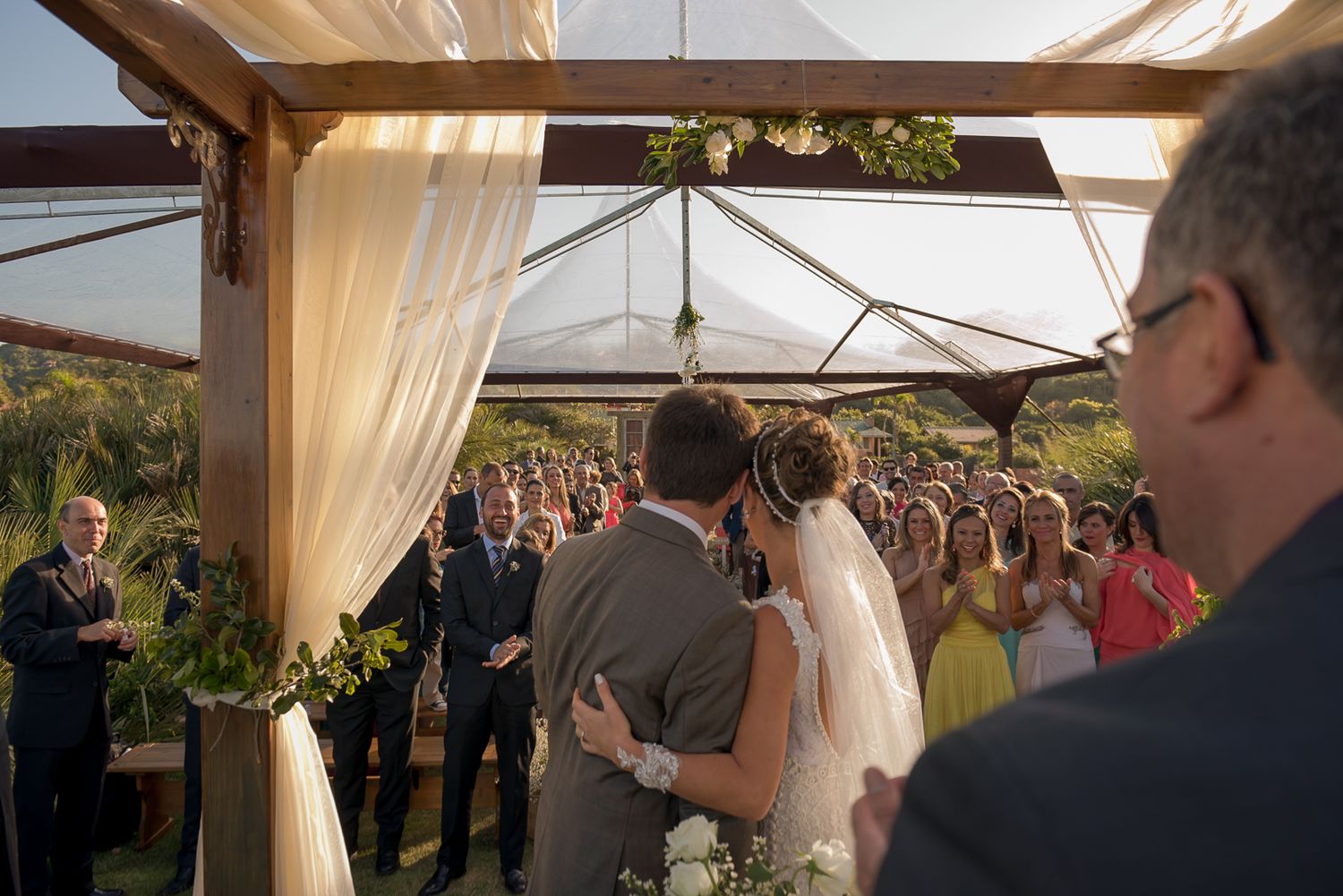 A wedding ceremony takes place under an elegant outdoor canopy with flowing white curtains and gathered guests.