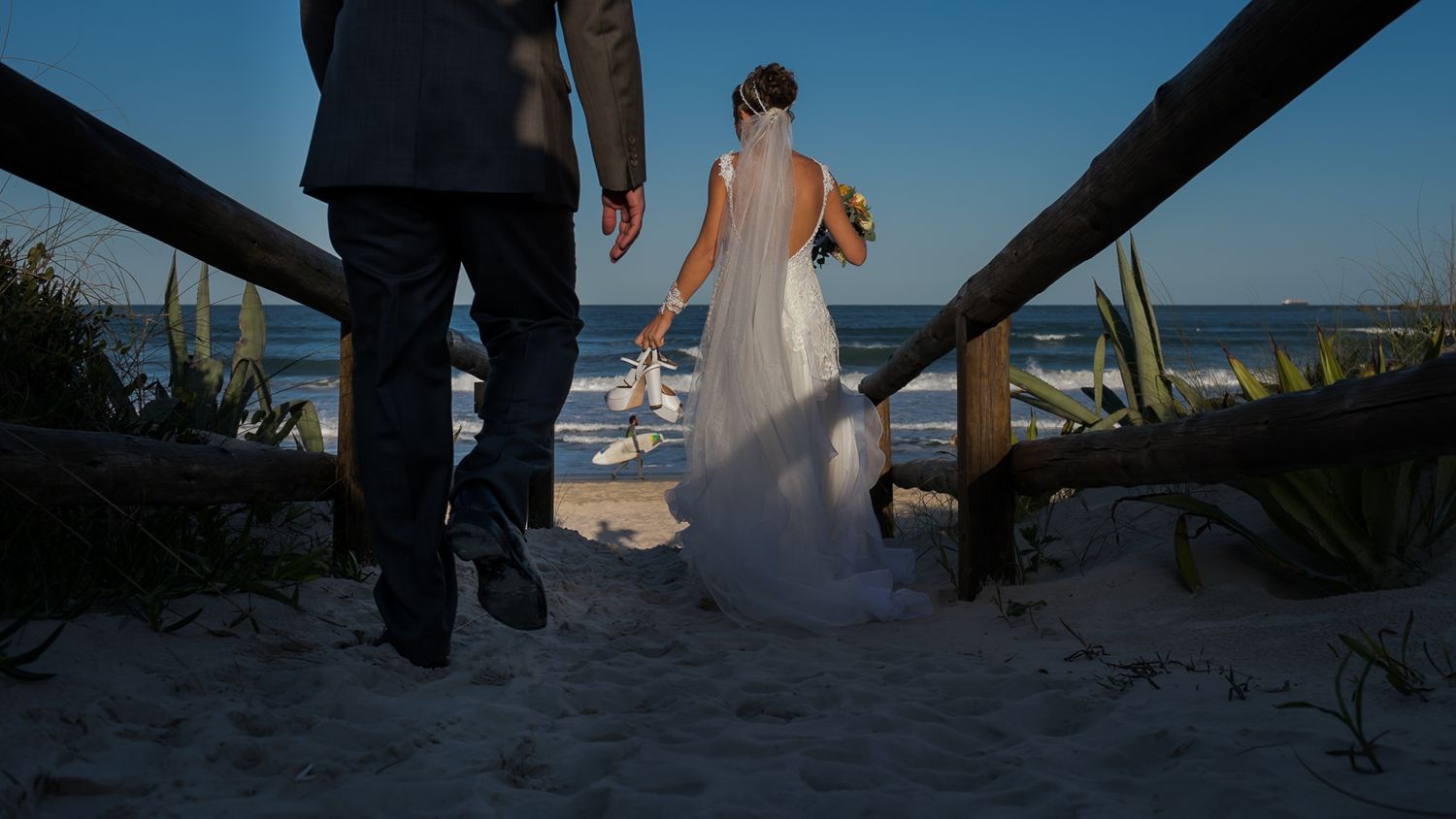 A couple and guest walk down wooden stairs to a beach at sunset, the bride's dress trailing behind.