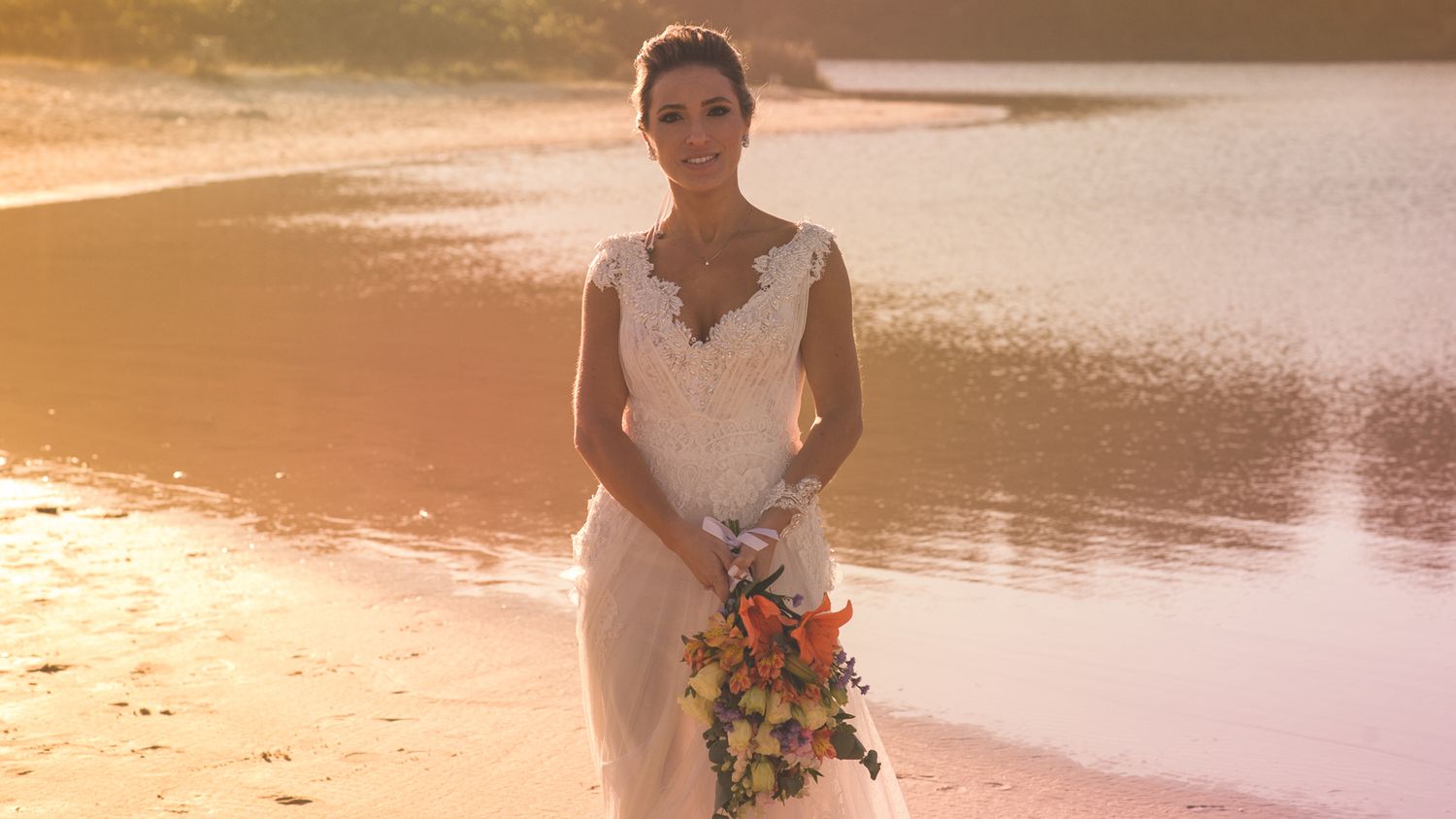A bride poses in a lace wedding dress holding a colorful bouquet against a sunset lake backdrop.