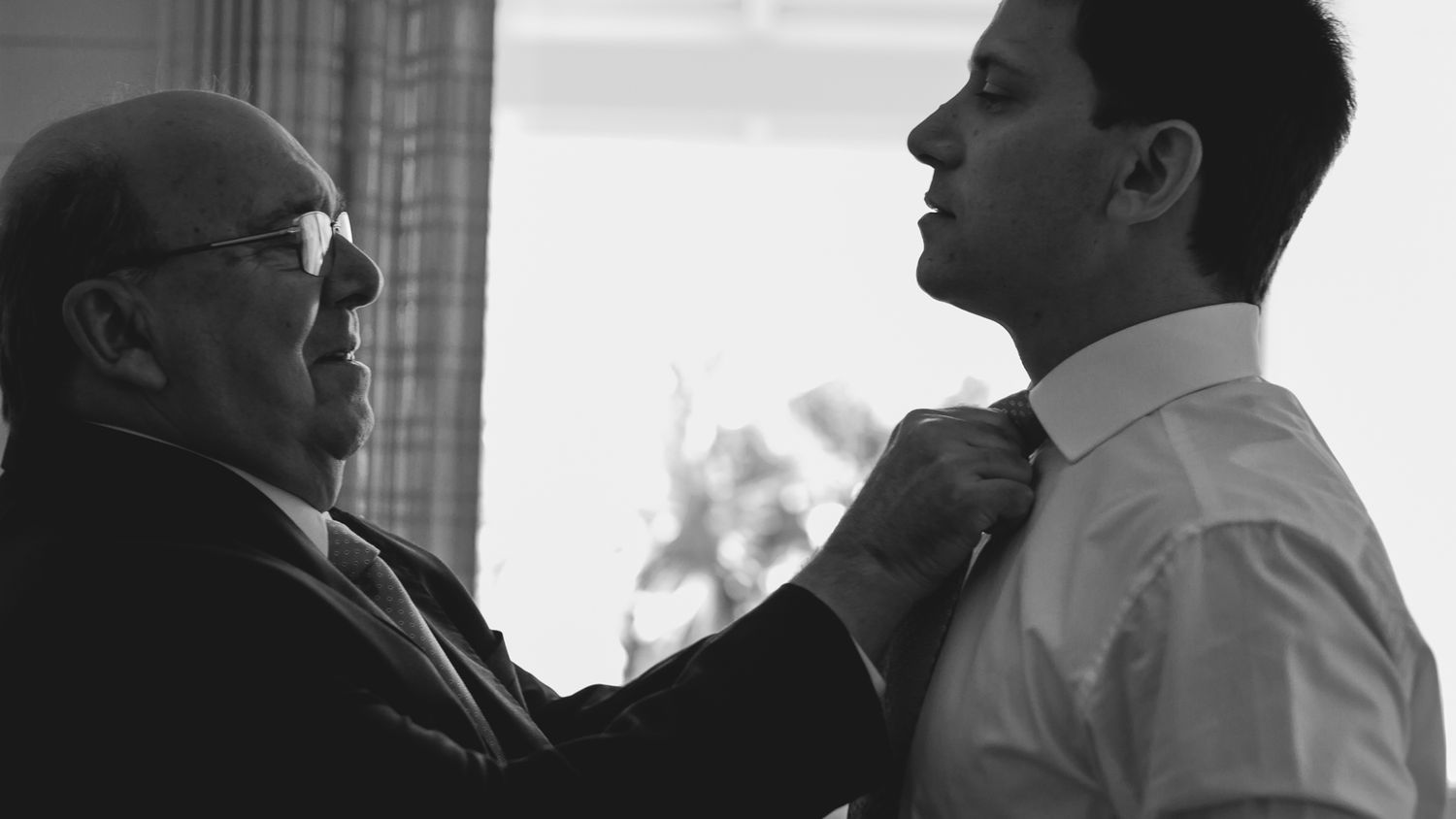 A black and white silhouette of two people during a formal dressing moment.
