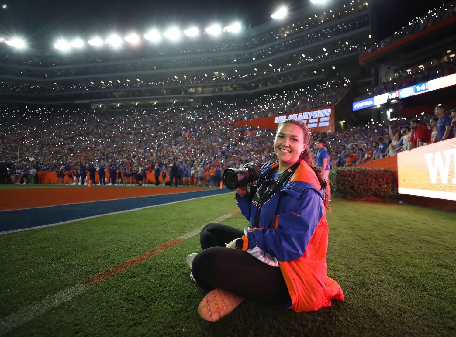 Person sitting on football field sideline at night with stadium lights and crowd in background.