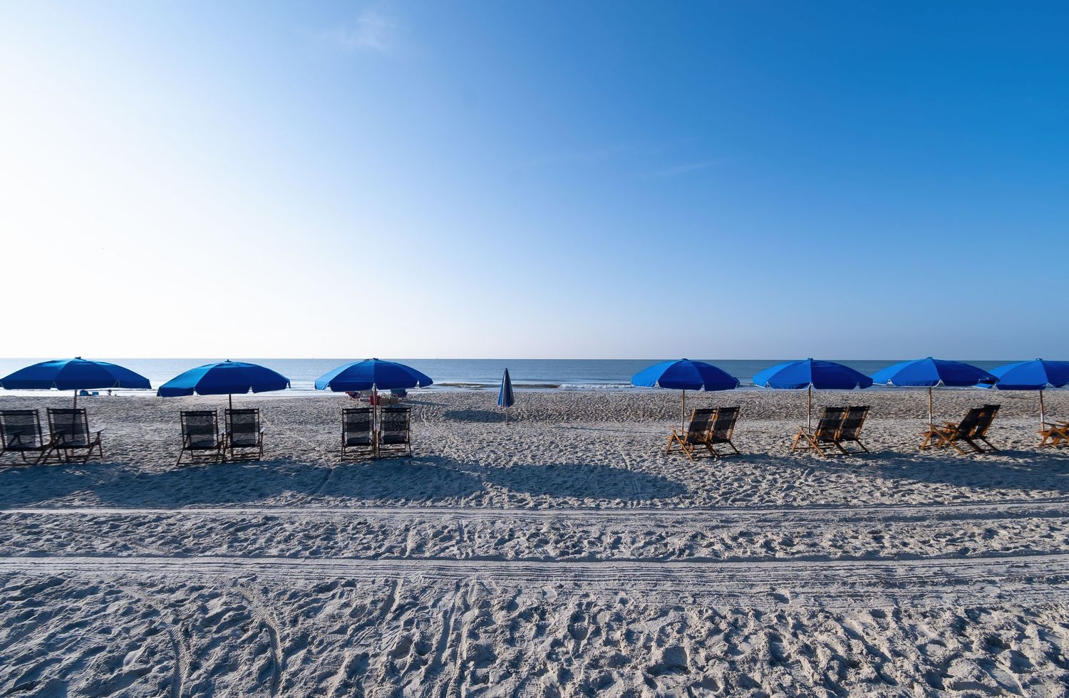Row of blue beach umbrellas and lounge chairs on empty sandy beach.