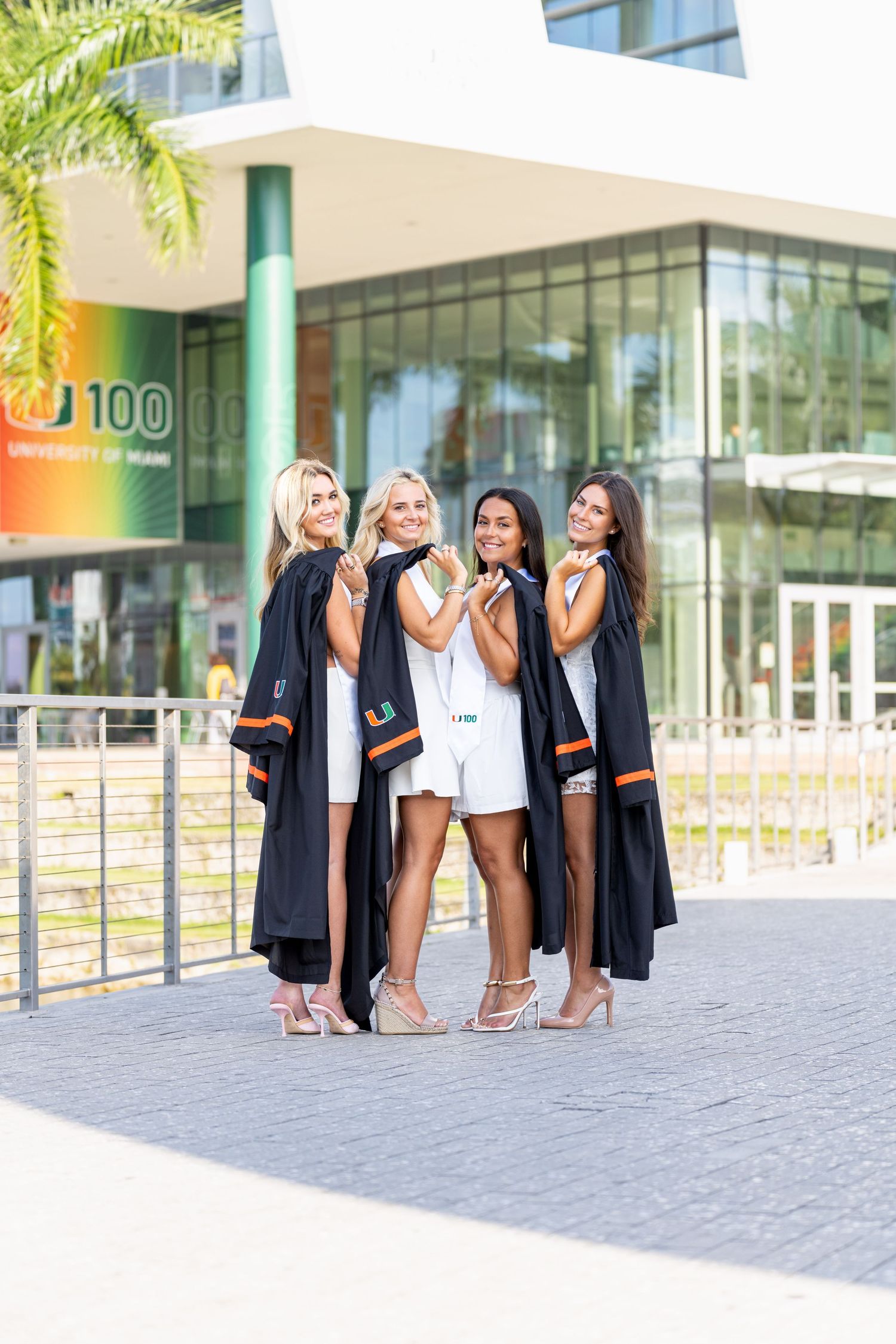 A group wearing graduation gowns poses for photos in front of a modern building with palm trees.