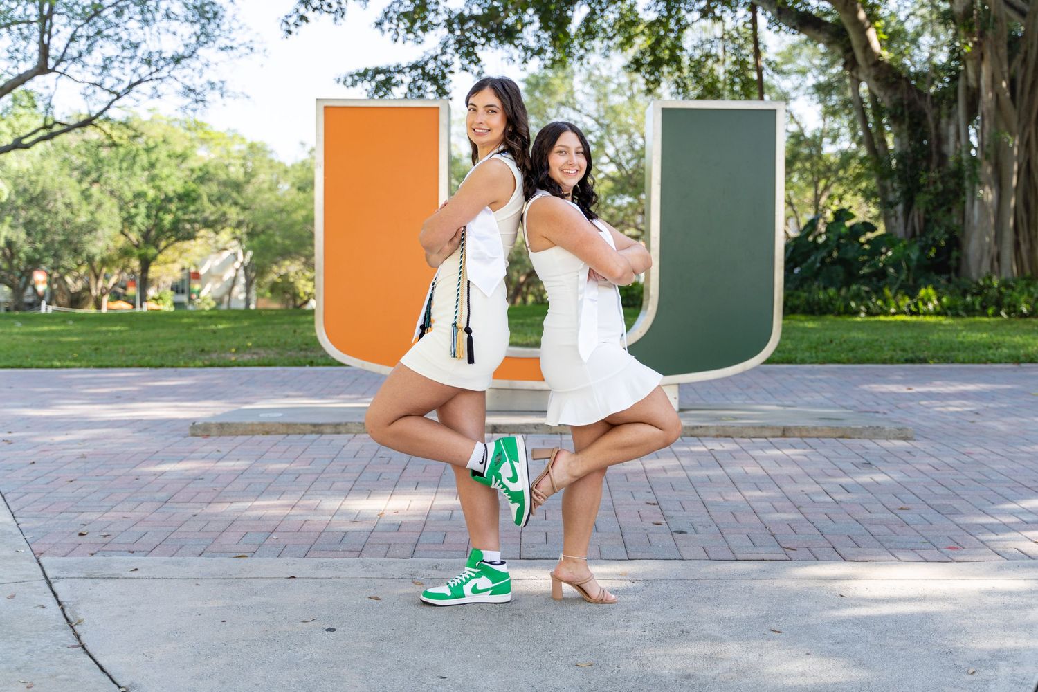 Students posing in graduation attire near outdoor branded university signage with orange and green U logos.