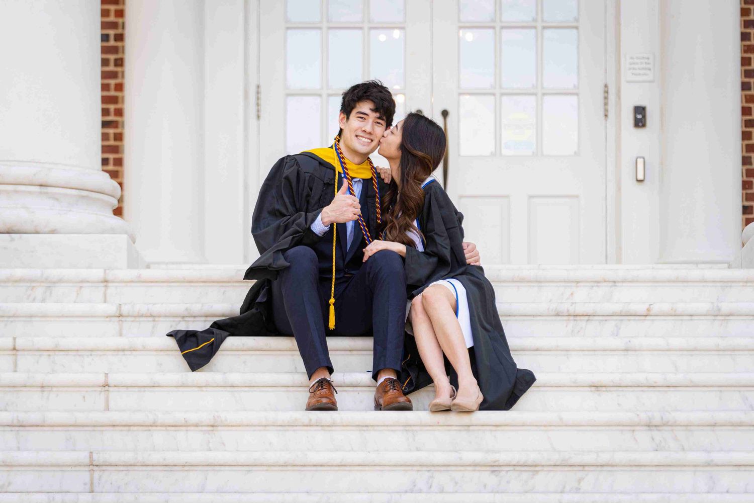 Two graduates in caps and gowns sitting close together on white stone steps.