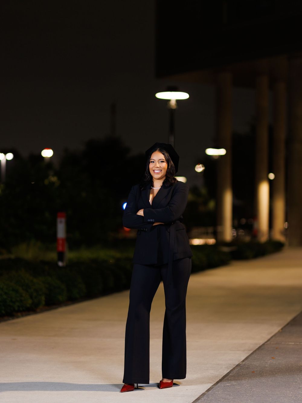 Woman in black pantsuit and red heels stands confidently with arms crossed on a walkway at night.