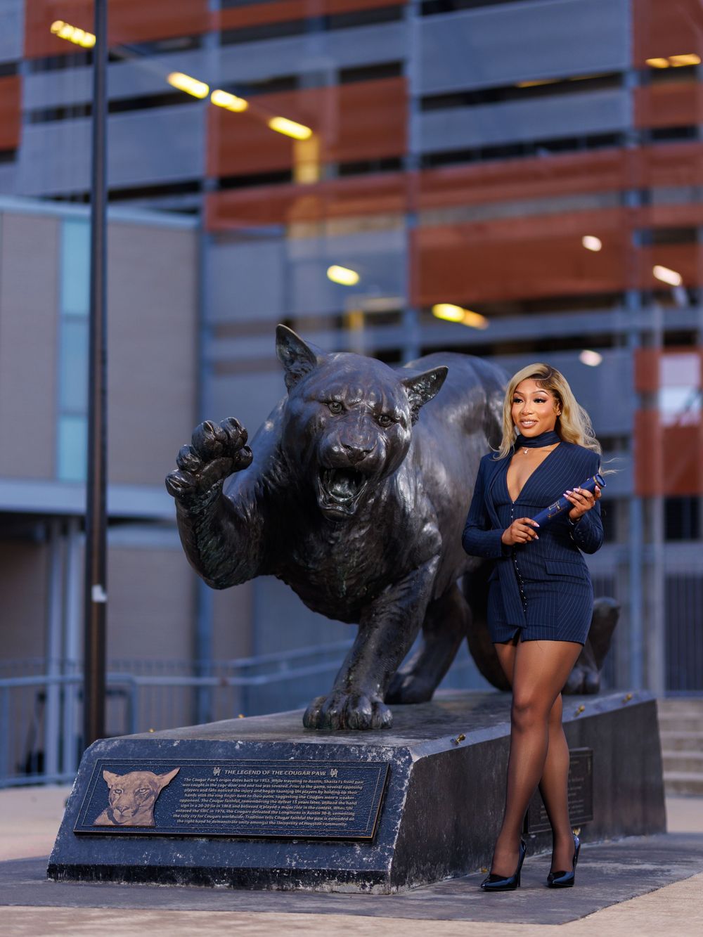 Woman in navy blue romper posing beside a bronze panther statue outside a modern building at night.