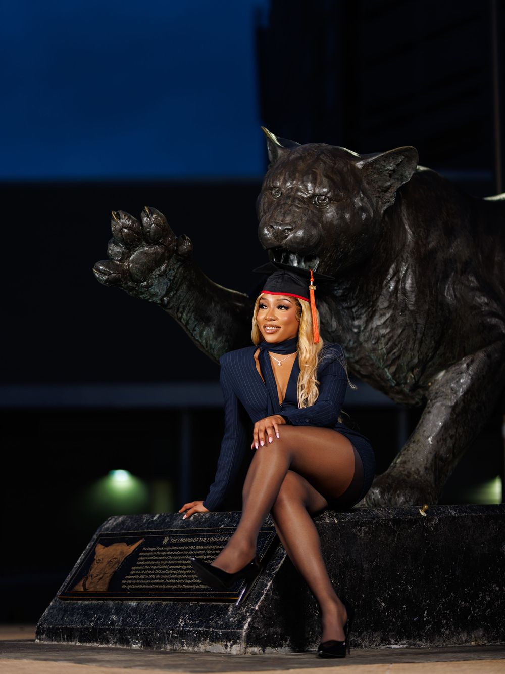 Graduate in cap and gown poses confidently beside a large bronze panther statue at night on college campus.