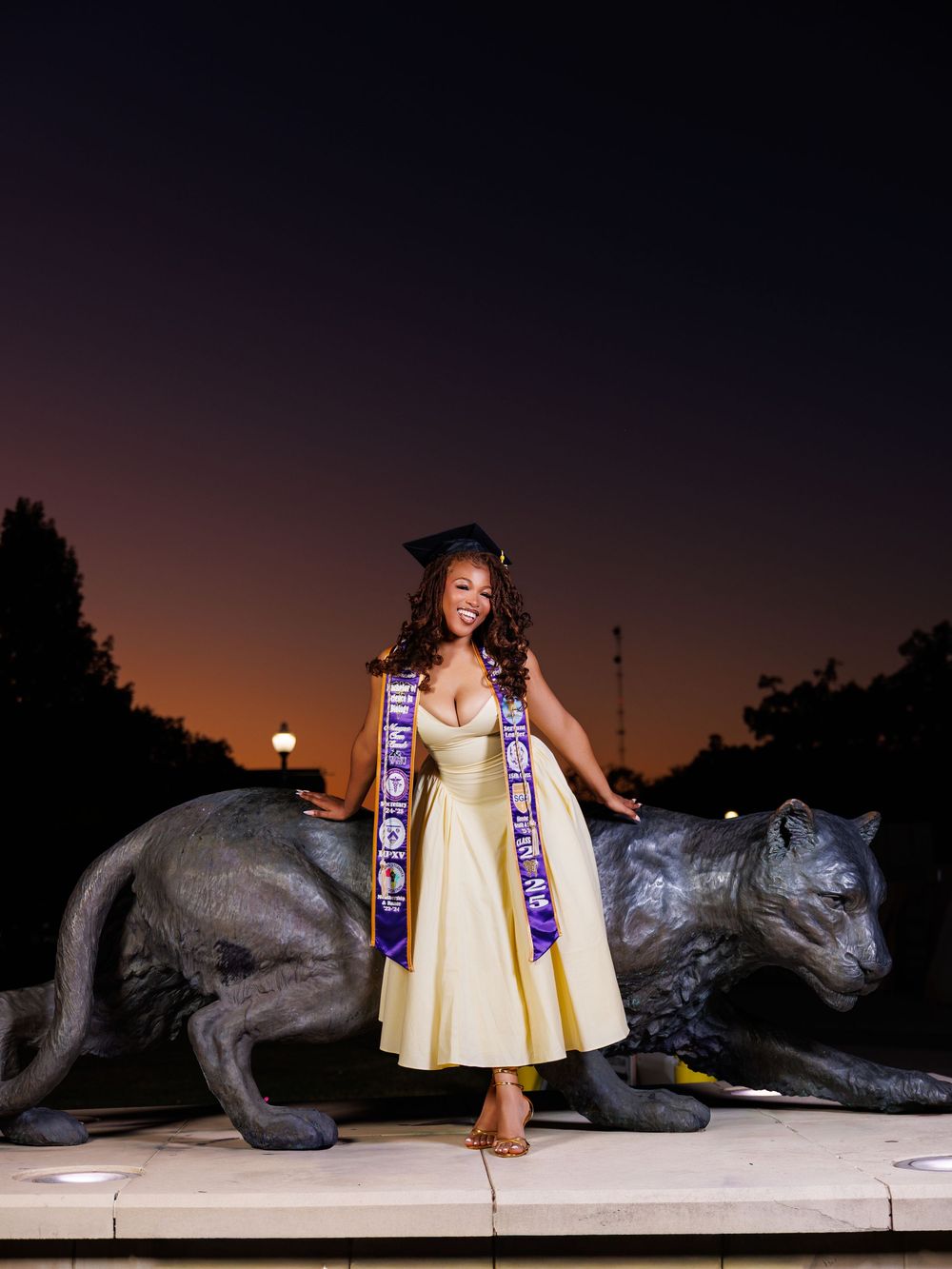 A person in a yellow dress poses with LSU tiger mascot statues at sunset on campus.