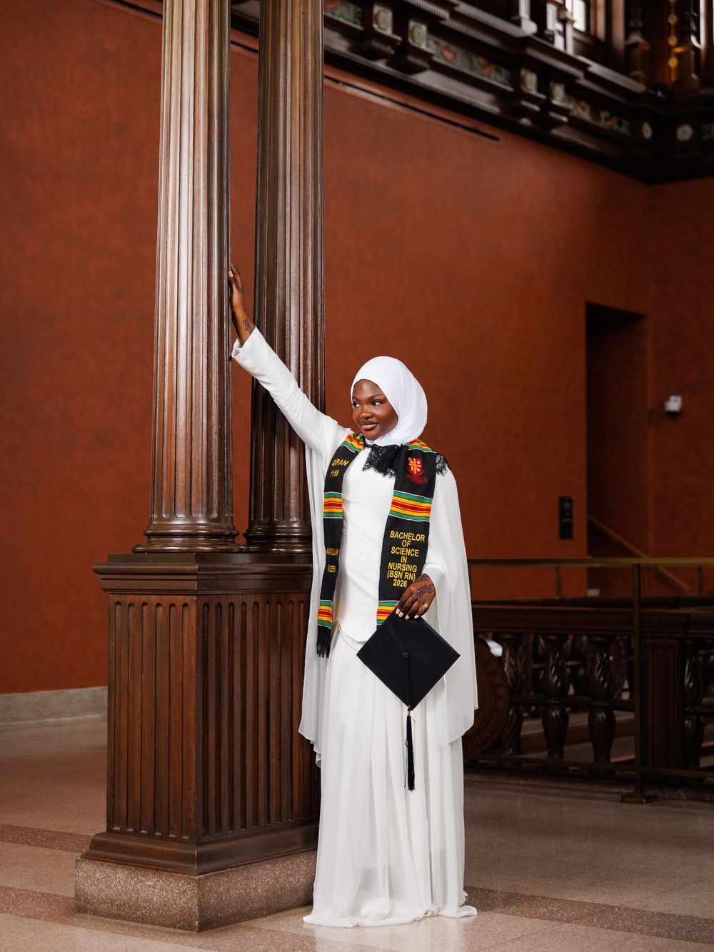 Graduate in white dress and kente stole holds cap beside ornate column in elegant hall.