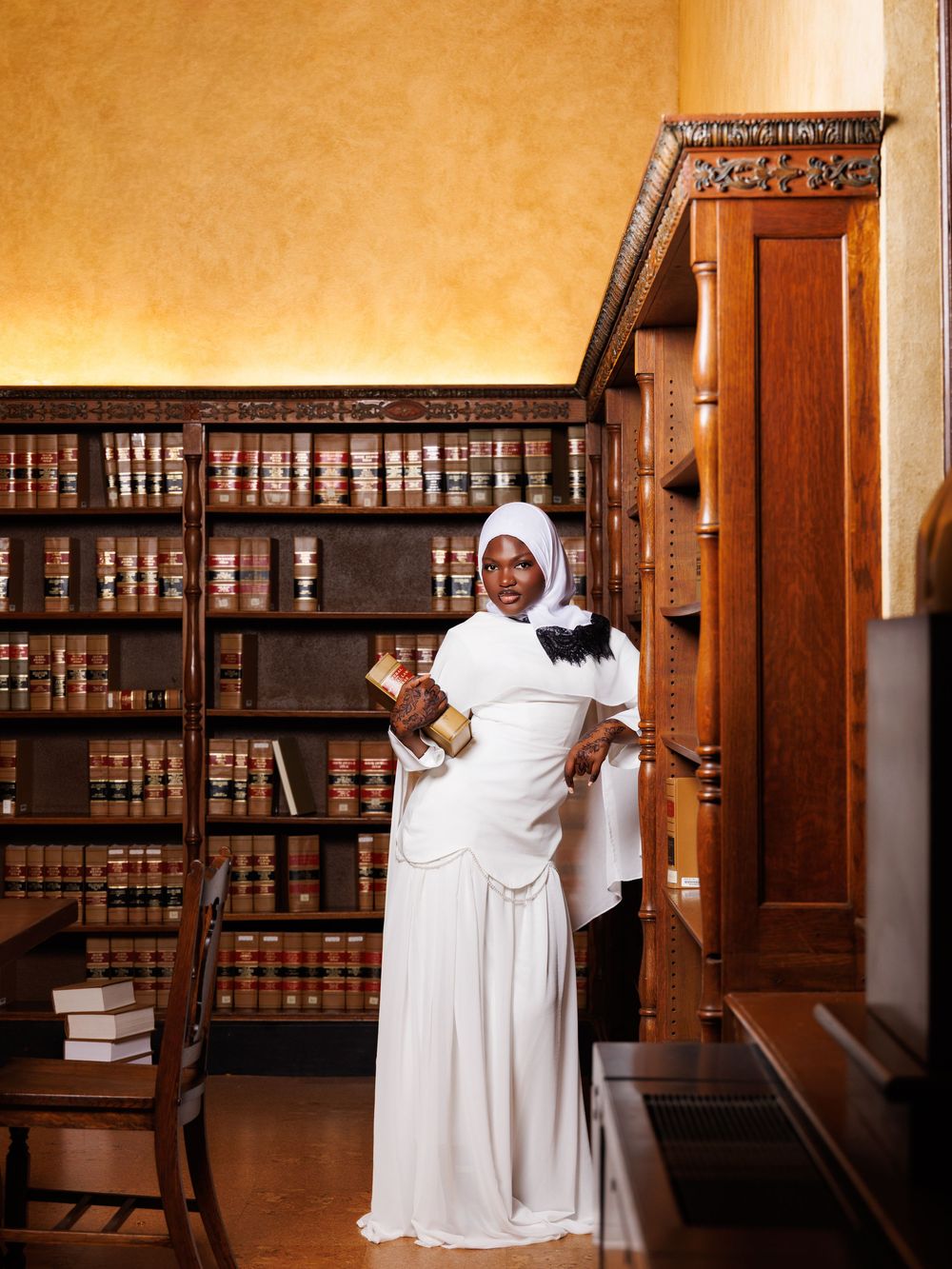 Woman in white dress and hijab holding books in an elegant wood-paneled library with shelves of leather-bound books.