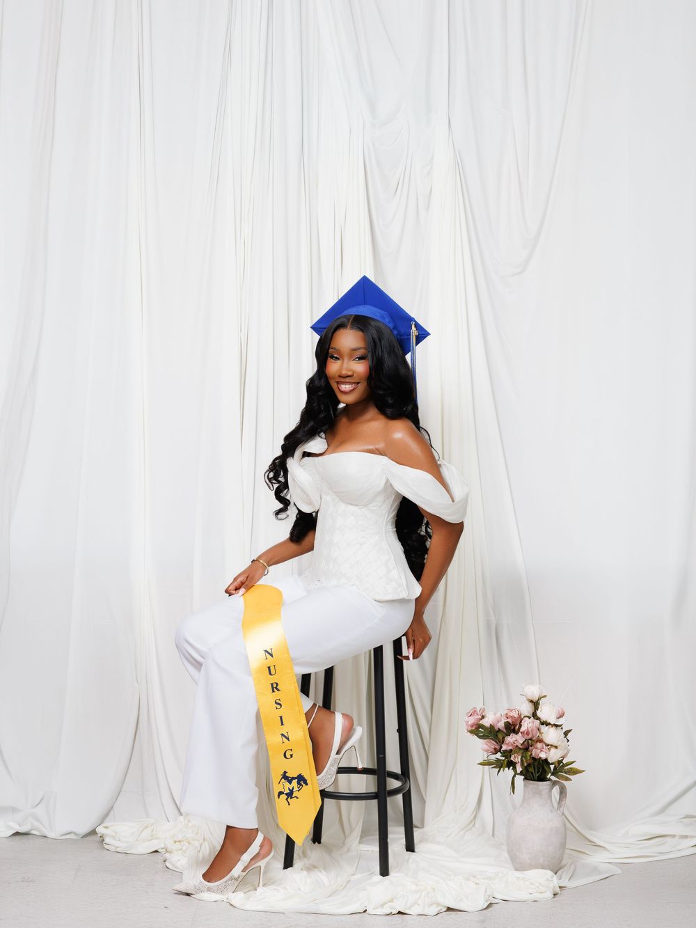 A young woman in a white outfit and blue graduation cap poses with a yellow sash on a stool against a white draped backdrop.