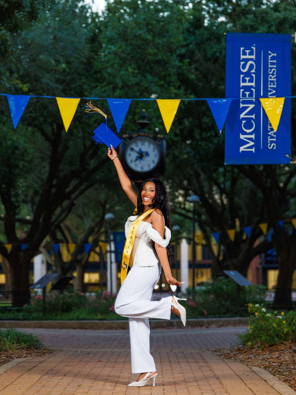 McNeese State University graduate in white outfit celebrates outdoors with cap, gold sash, and blue-yellow pennant banners.