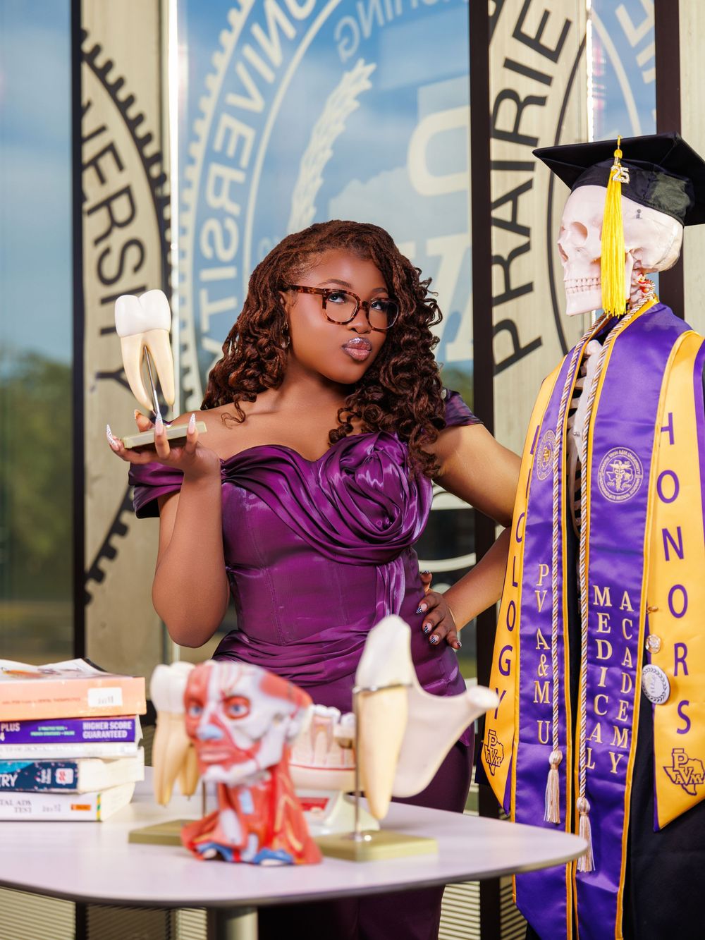 Student in purple dress poses with academic awards and banners at graduation celebration event.