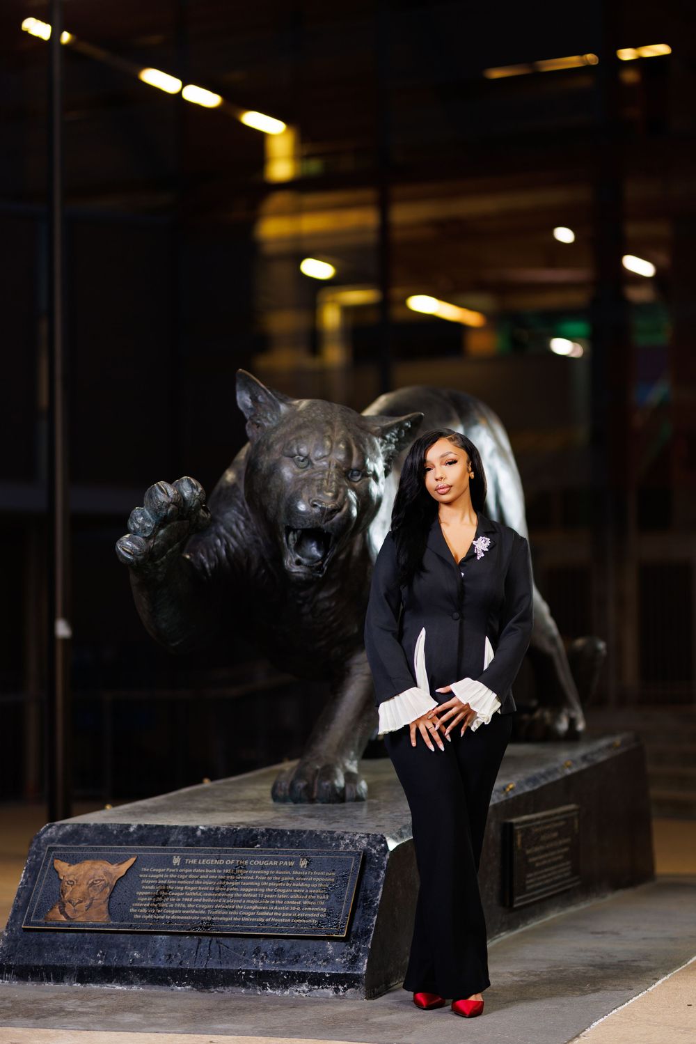 A person in a black outfit poses with a large bronze panther statue at night with illuminated lighting in the background.