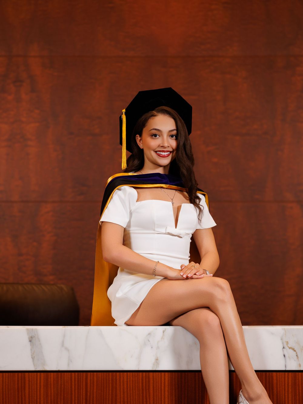 Woman in white dress and graduation cap and hood smiles while seated on marble surface at ceremony.