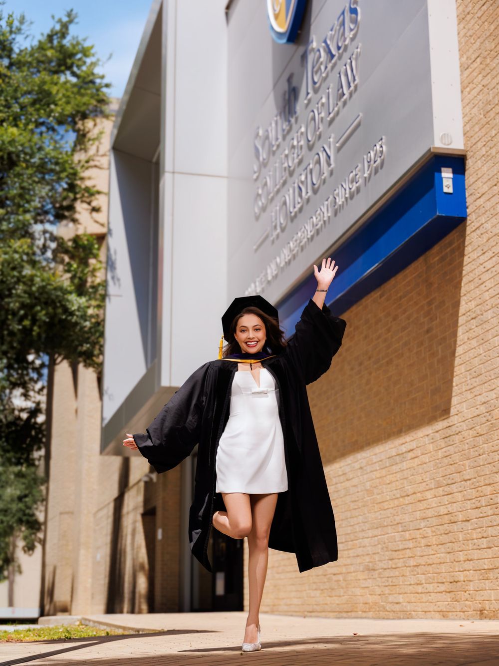 Smiling graduate in black cap and gown over white dress celebrates outdoors at university building.