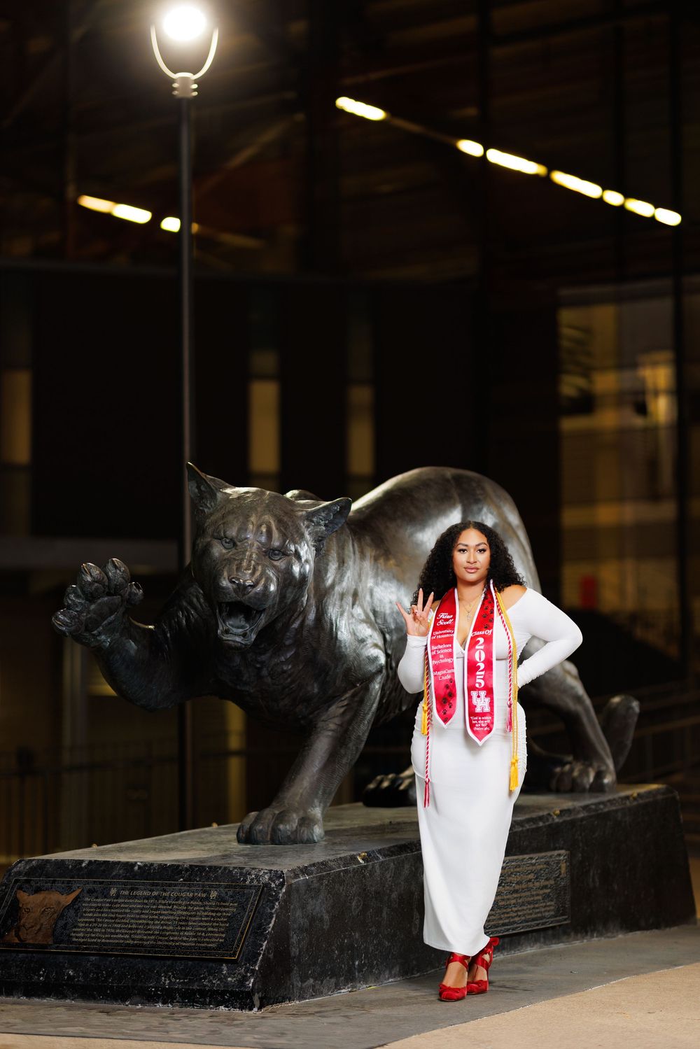 A person in a white dress and red shoes poses with a large bronze bull statue in a dark indoor space.