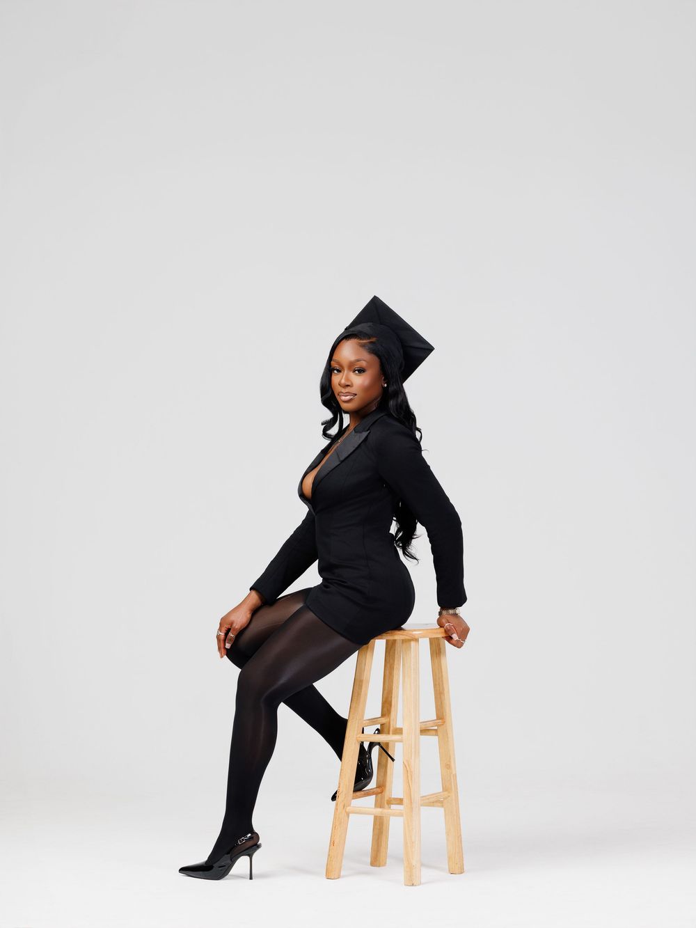 A person in black graduation attire sits on a wooden stool in various poses against a white studio backdrop.