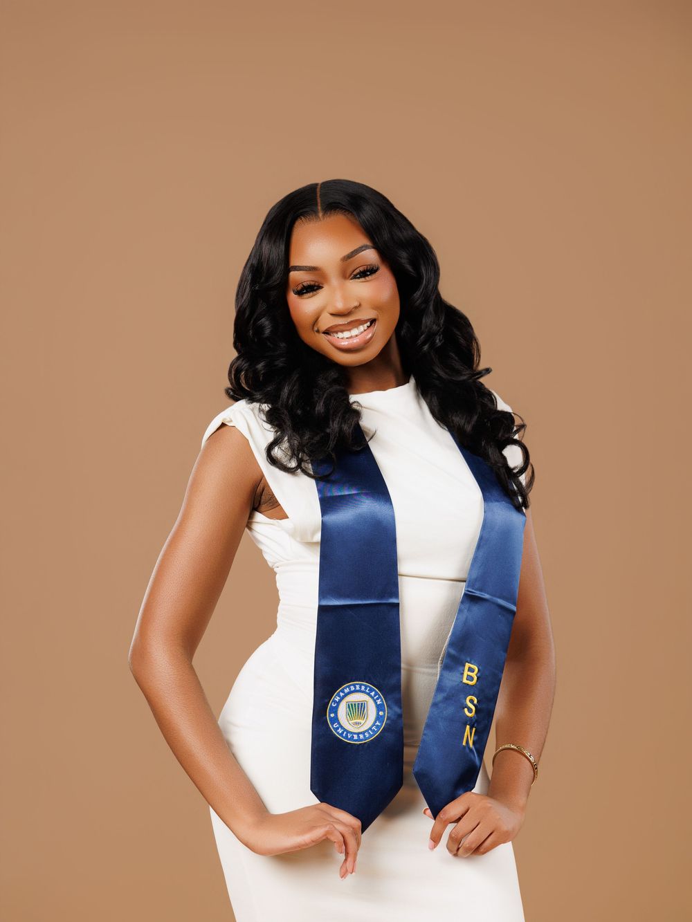 Graduate wearing white dress and blue graduation sash poses for portrait against brown backdrop.