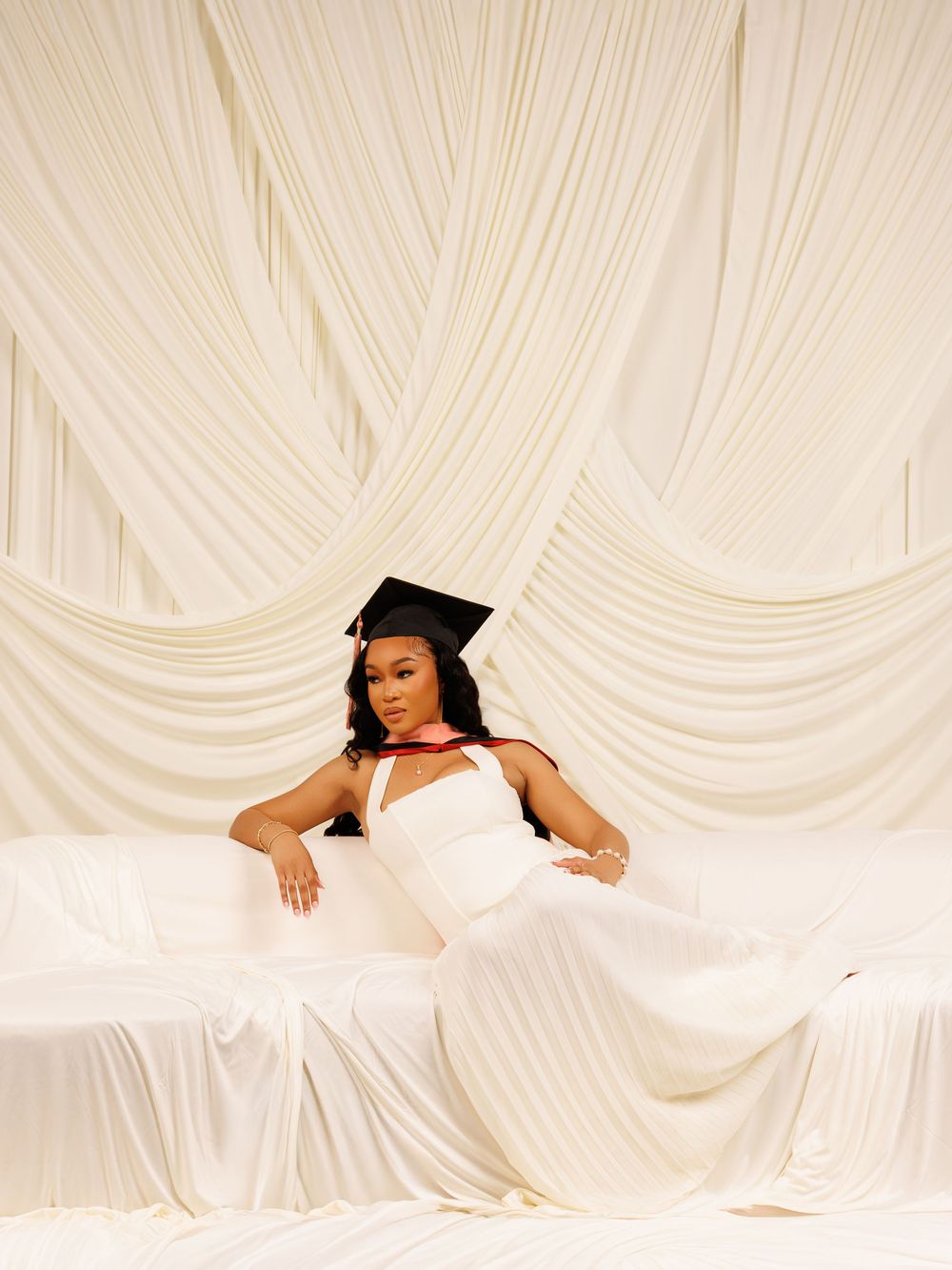 Woman in white dress and graduation cap poses elegantly on white draped couch against cream fabric backdrop.