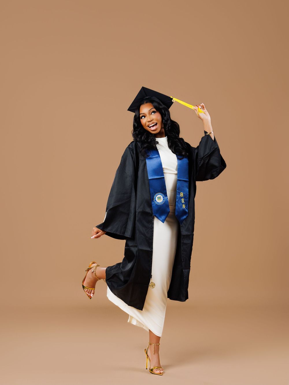 Graduate in black cap and gown with blue stole poses playfully on brown background during graduation photoshoot.