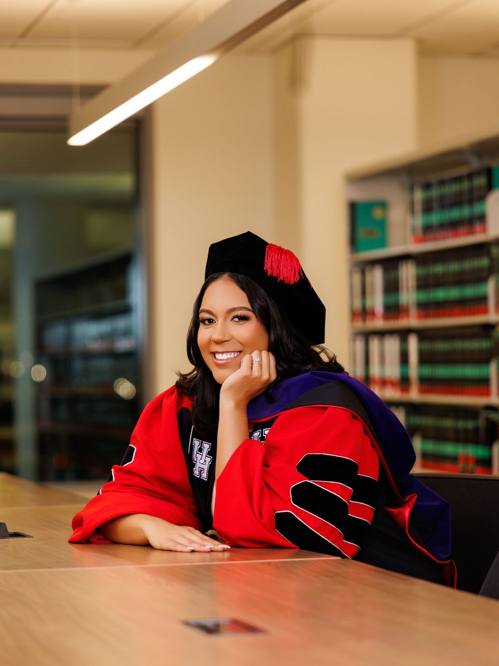 Woman in red doctoral graduation regalia and cap smiles proudly while seated in a university library.