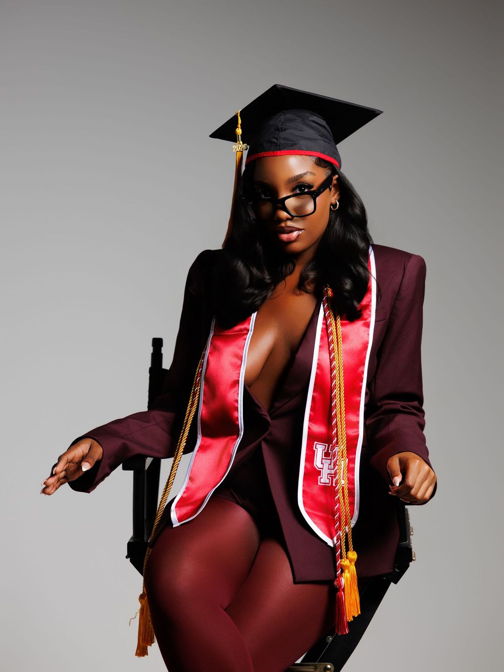 A young woman in graduation cap, red sash, and maroon blazer poses confidently in a studio setting.