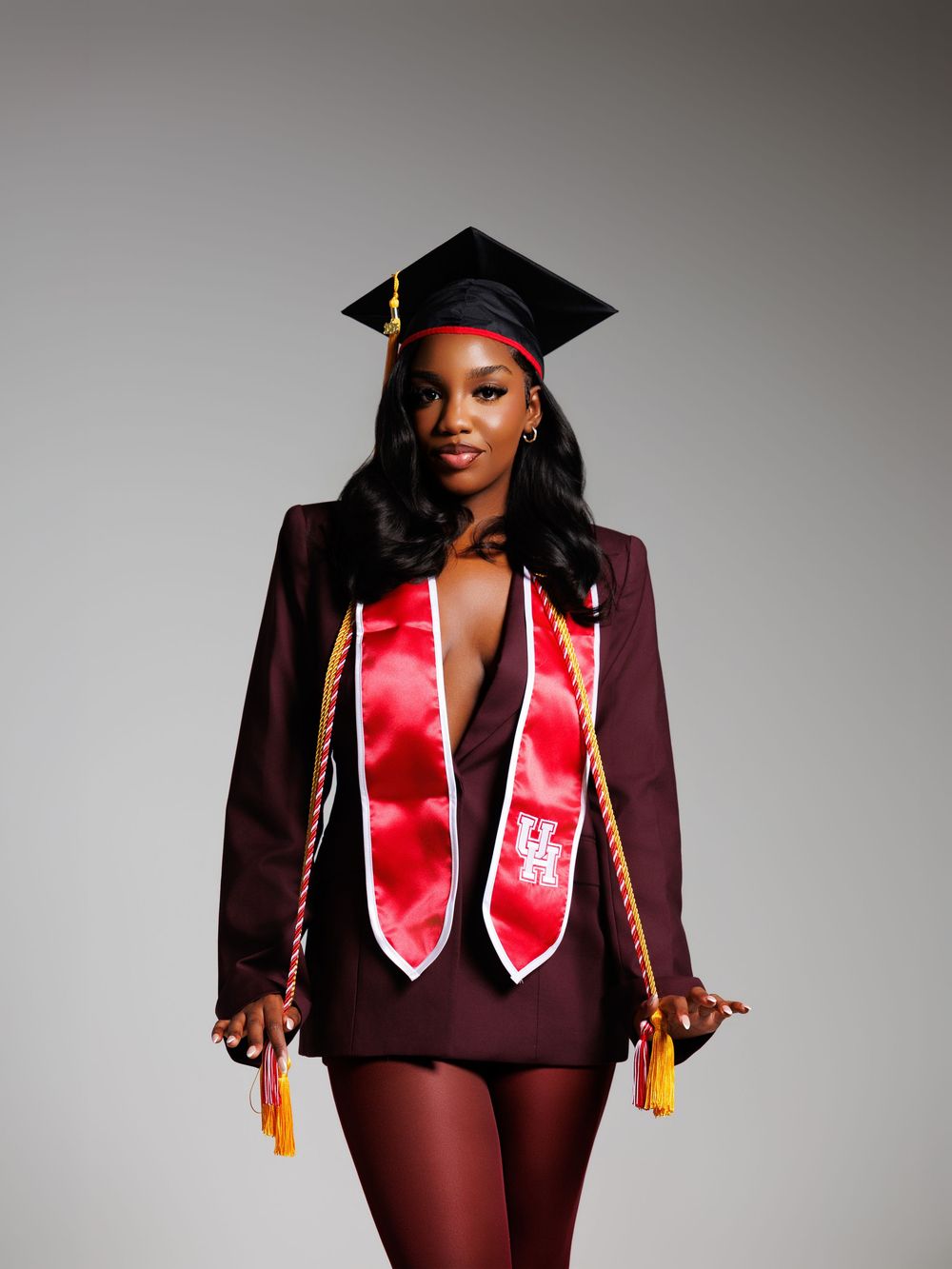 Woman in maroon graduation cap and gown with red honor stole and gold cords poses confidently for grad photos.