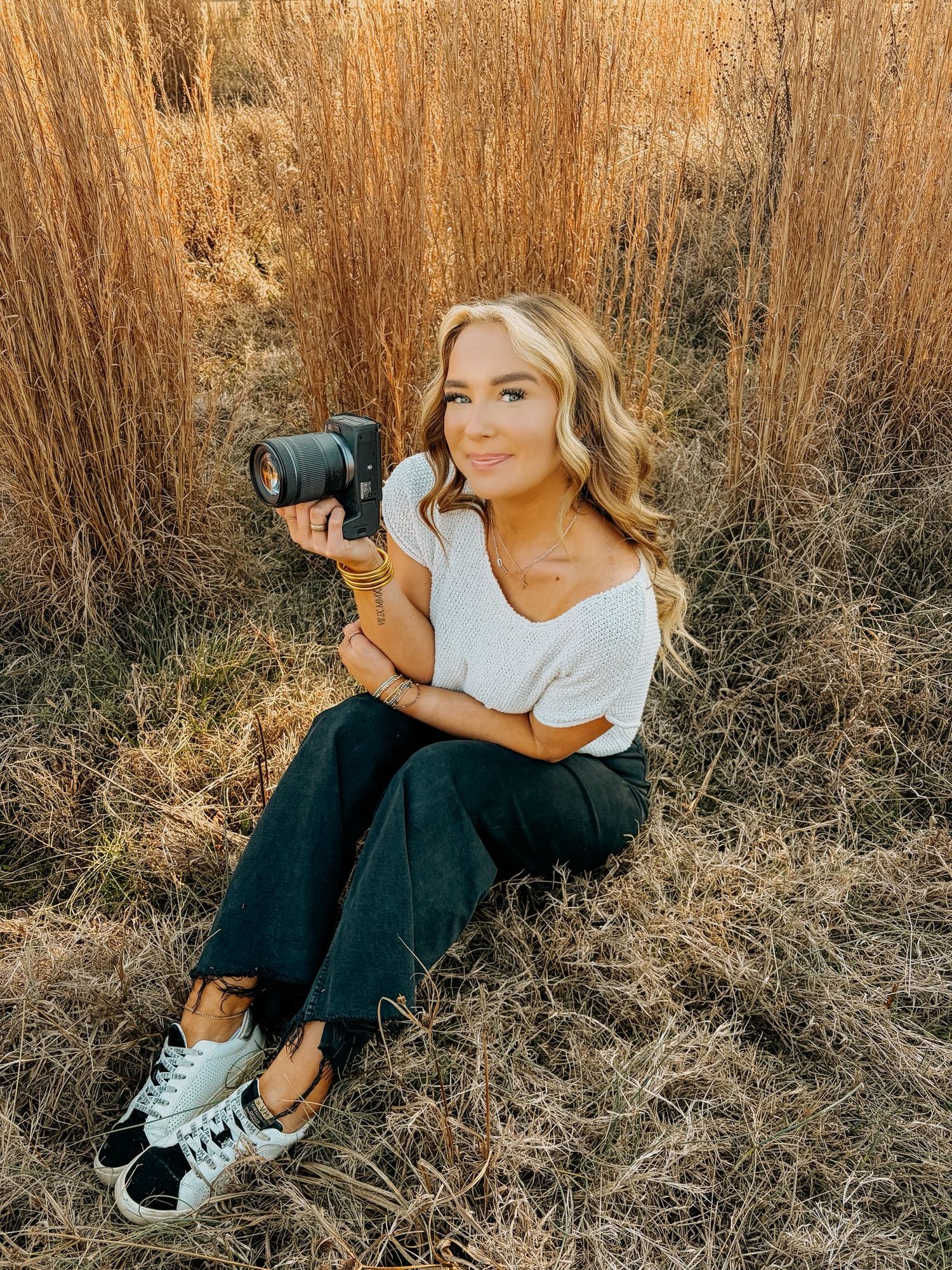 Photographer with long hair in coral sweater poses with camera in field of tall wheat grass.