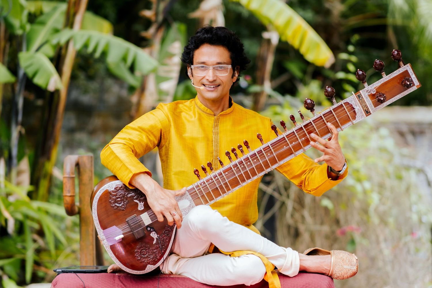 Musician in yellow traditional attire plays sitar while sitting cross-legged outdoors with tropical foliage.