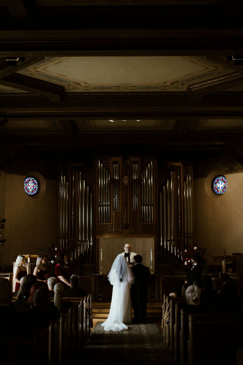 A dimly lit church sanctuary with rows of wooden pews and an organ pipe display illuminated by stained glass windows.