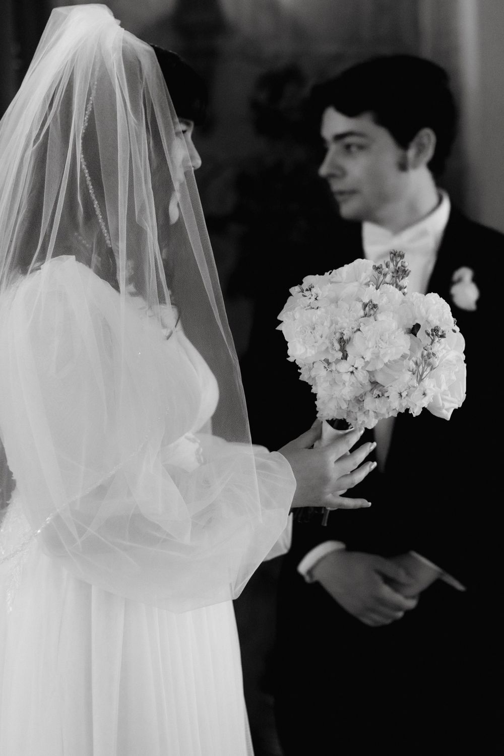 A black and white wedding photo showing the bride's white dress and veil with delicate floral bouquet details.