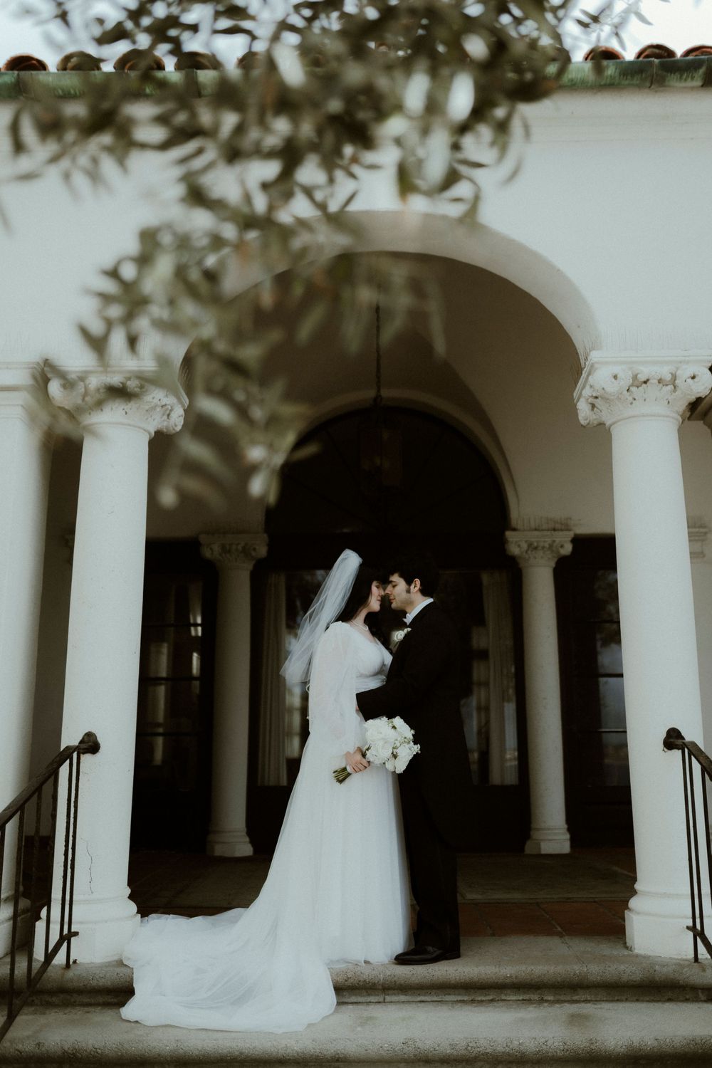 Wedding couple poses among white columns and arches in classic architectural setting.