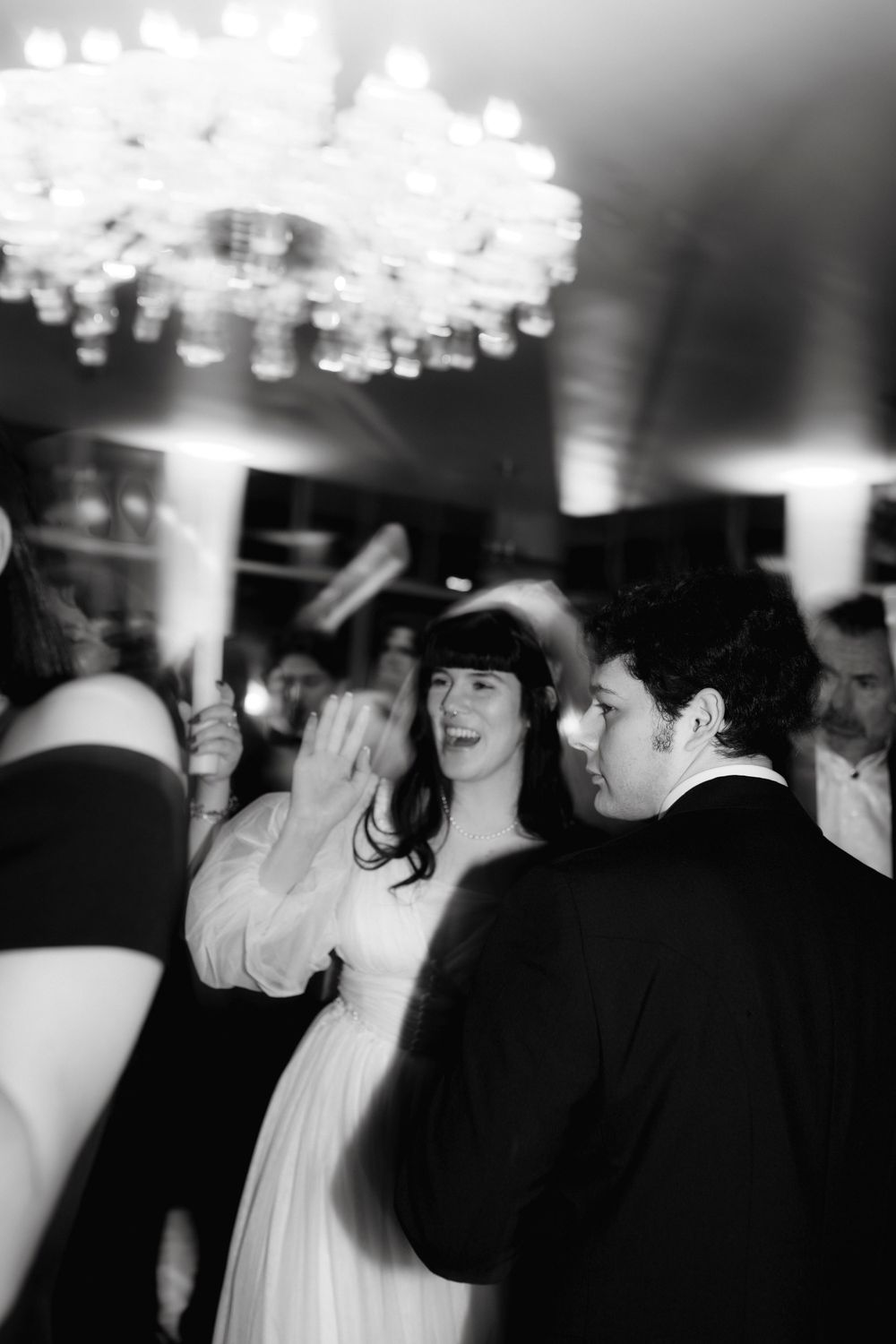 Blurred black and white image of guests dancing under crystal chandeliers at an elegant event.