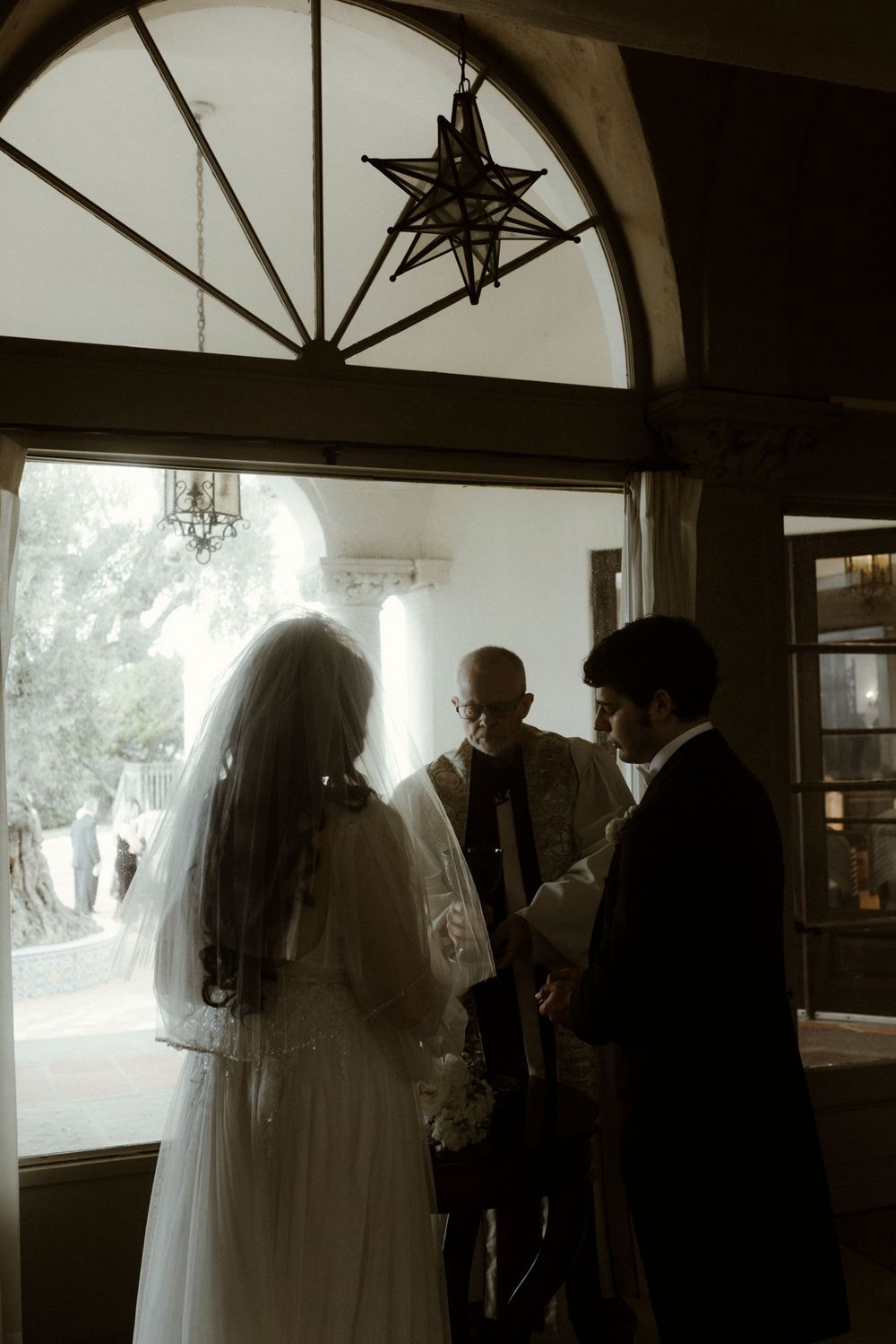Silhouettes of wedding guests and the bridal party stand in front of bright windows.