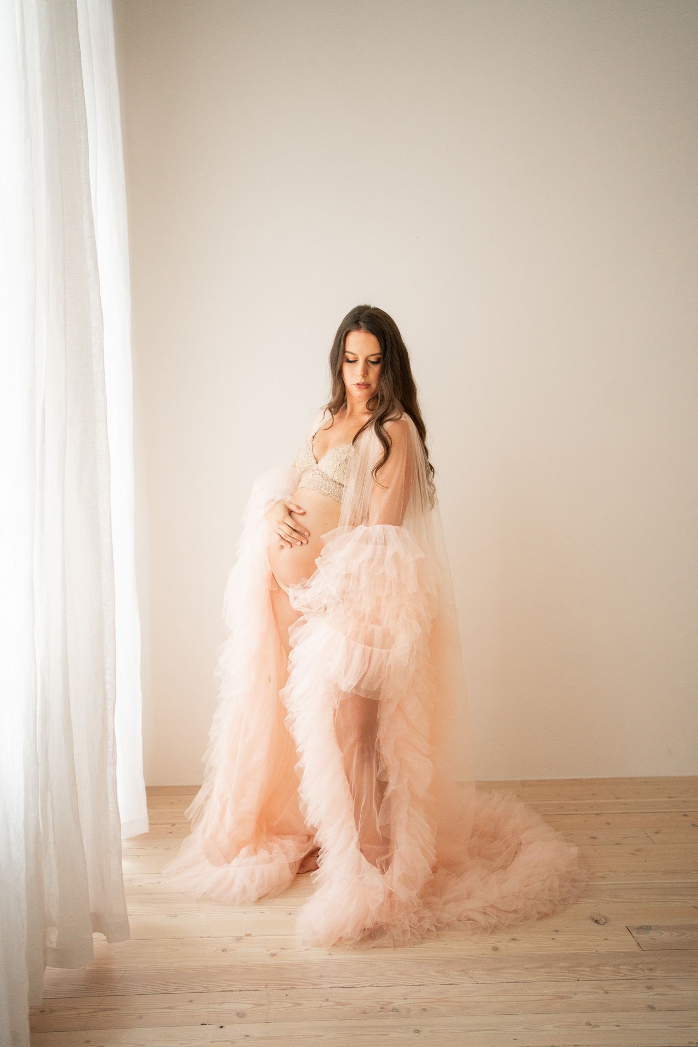 Ethereal maternity portrait in flowing pink tulle gown by bright window.