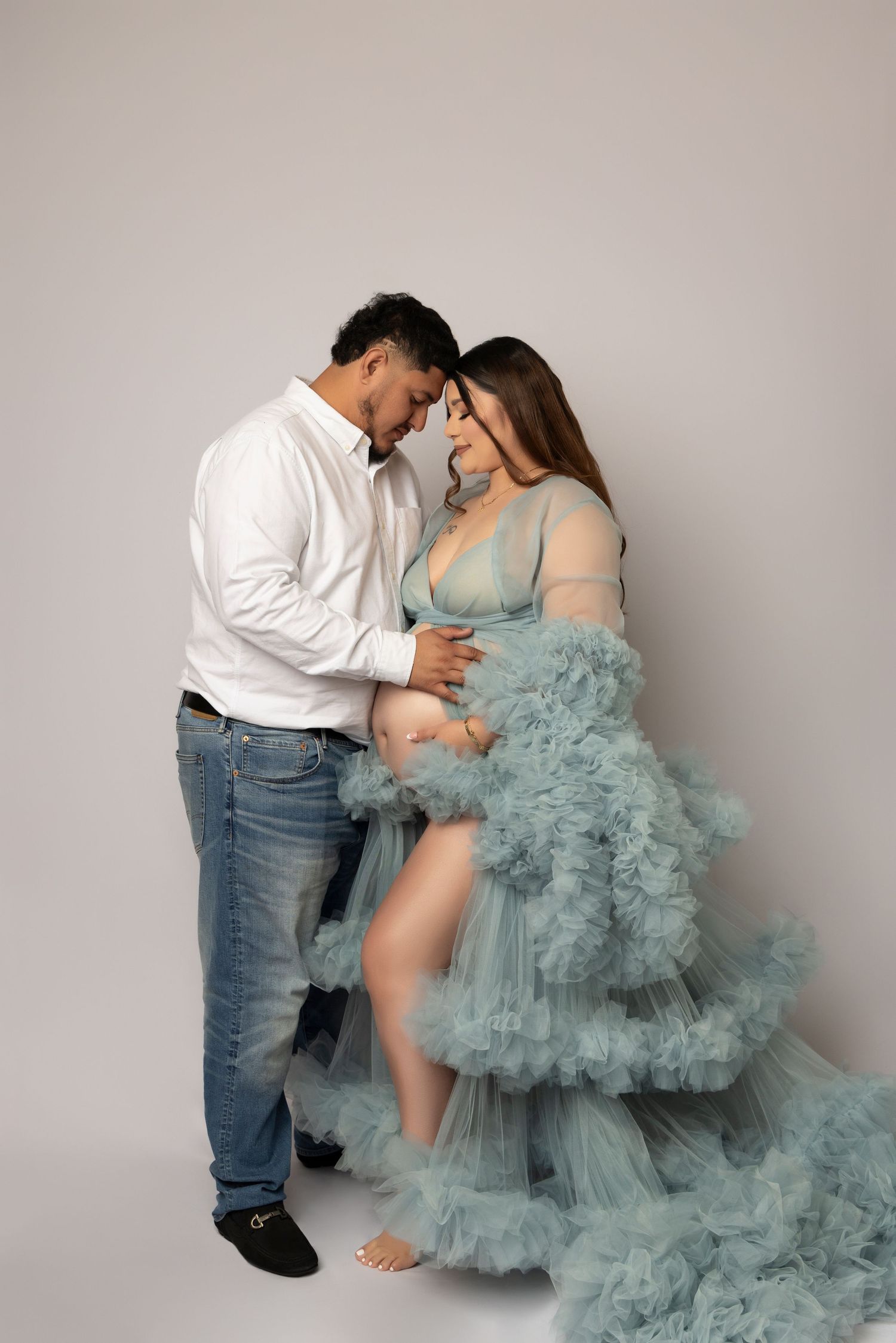 Maternity photo session with woman in flowing teal tulle gown against grey backdrop.